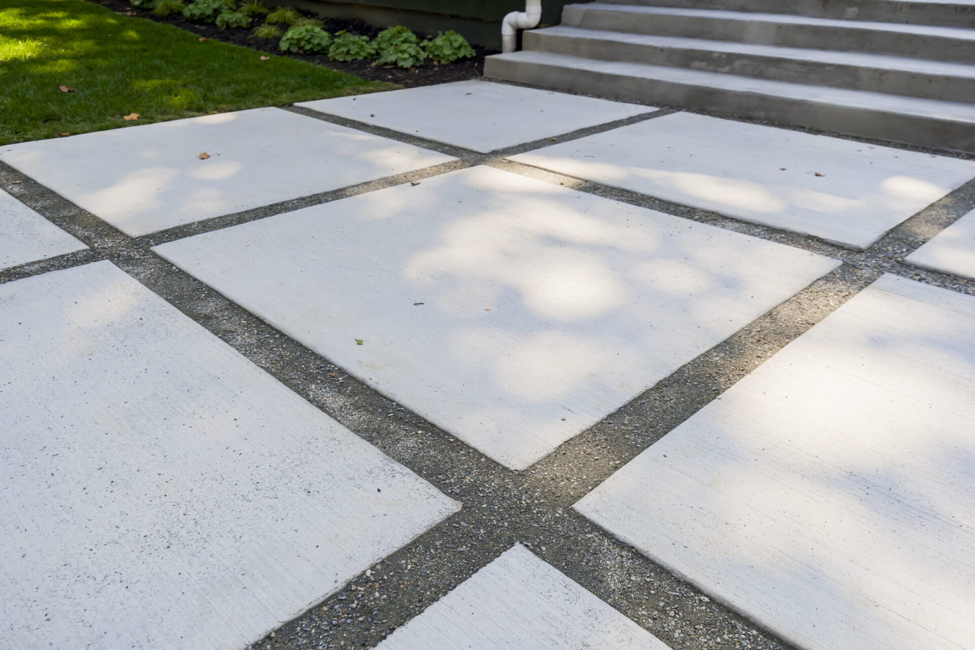 A concrete walkway with large square slabs bordered by small gravel, adjacent to a grassy area and leading to steps.