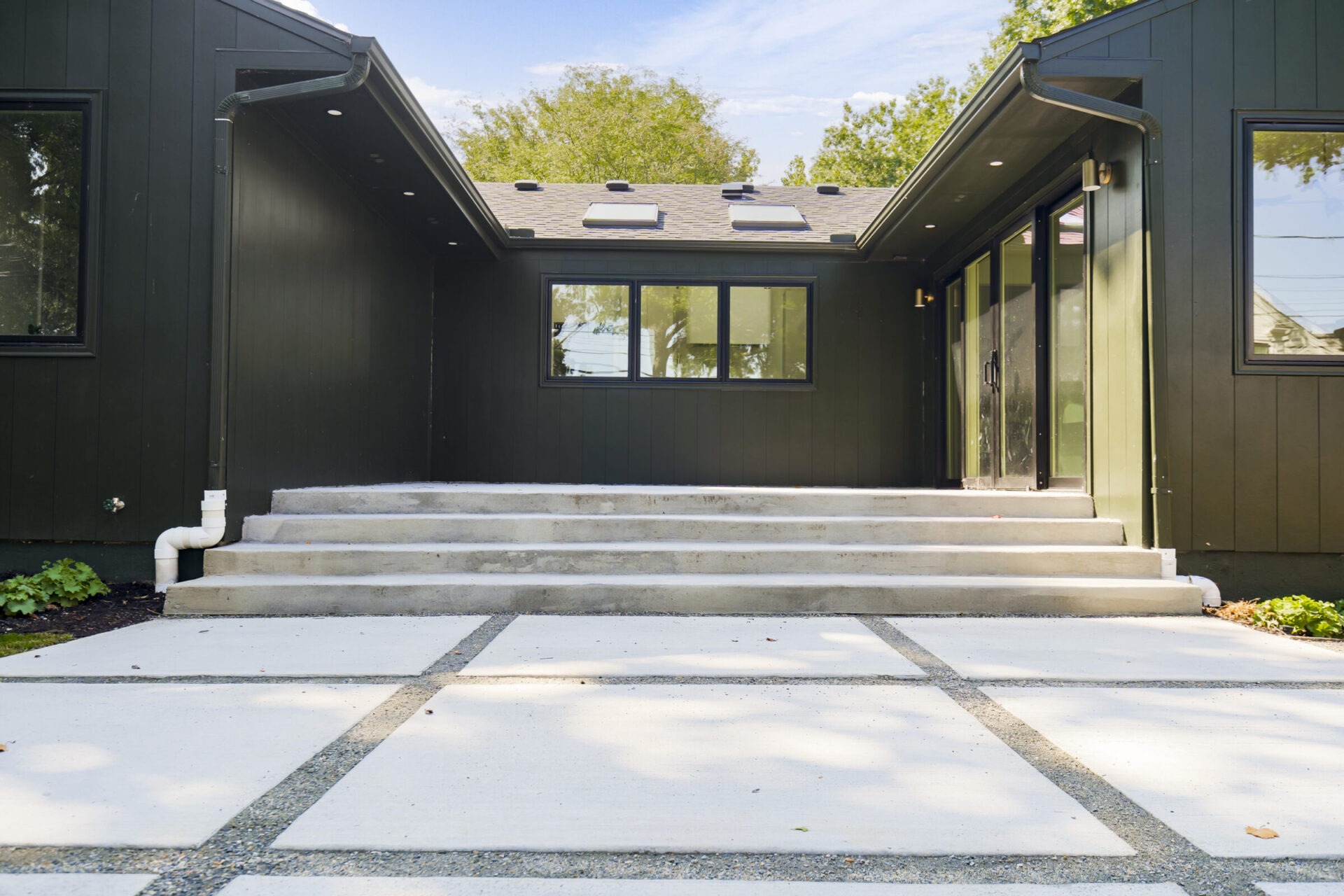 Modern, dark exterior with large windows and concrete steps, surrounded by greenery under a blue sky. No recognizable landmarks or historical buildings.