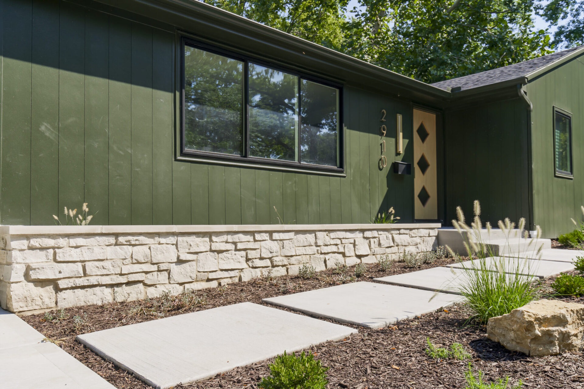 Green house exterior with large window, stone foundation, landscaped path, and a door featuring geometric design. Surrounded by greenery under sunny skies.