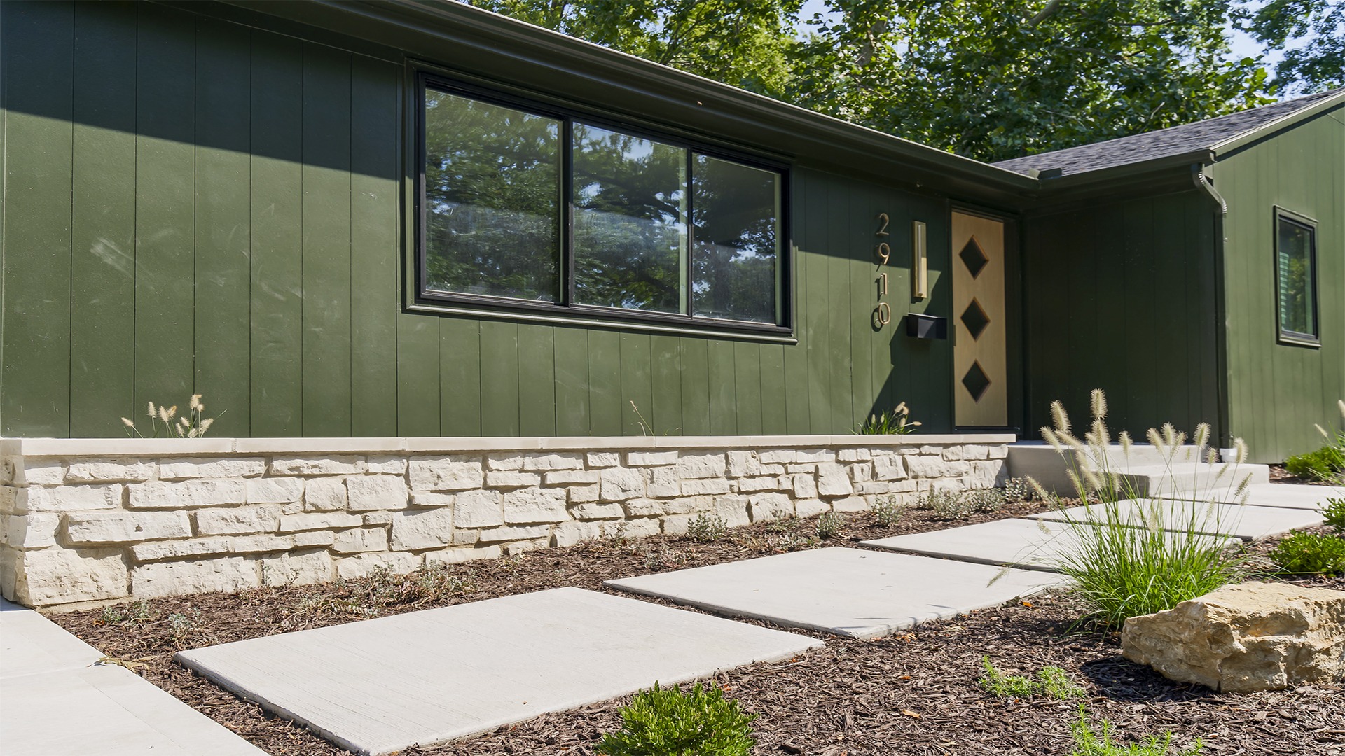 Green house exterior with stone accents, modern door, and landscaped path with greenery. Sunlight casts soft shadows on the facade.