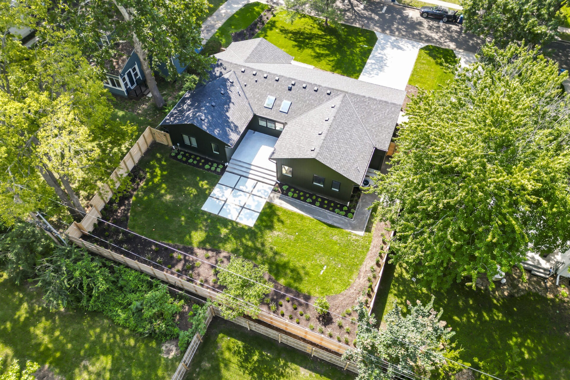 Aerial view of a modern house with a dark roof, surrounded by greenery and a wooden fence, featuring a spacious patio.