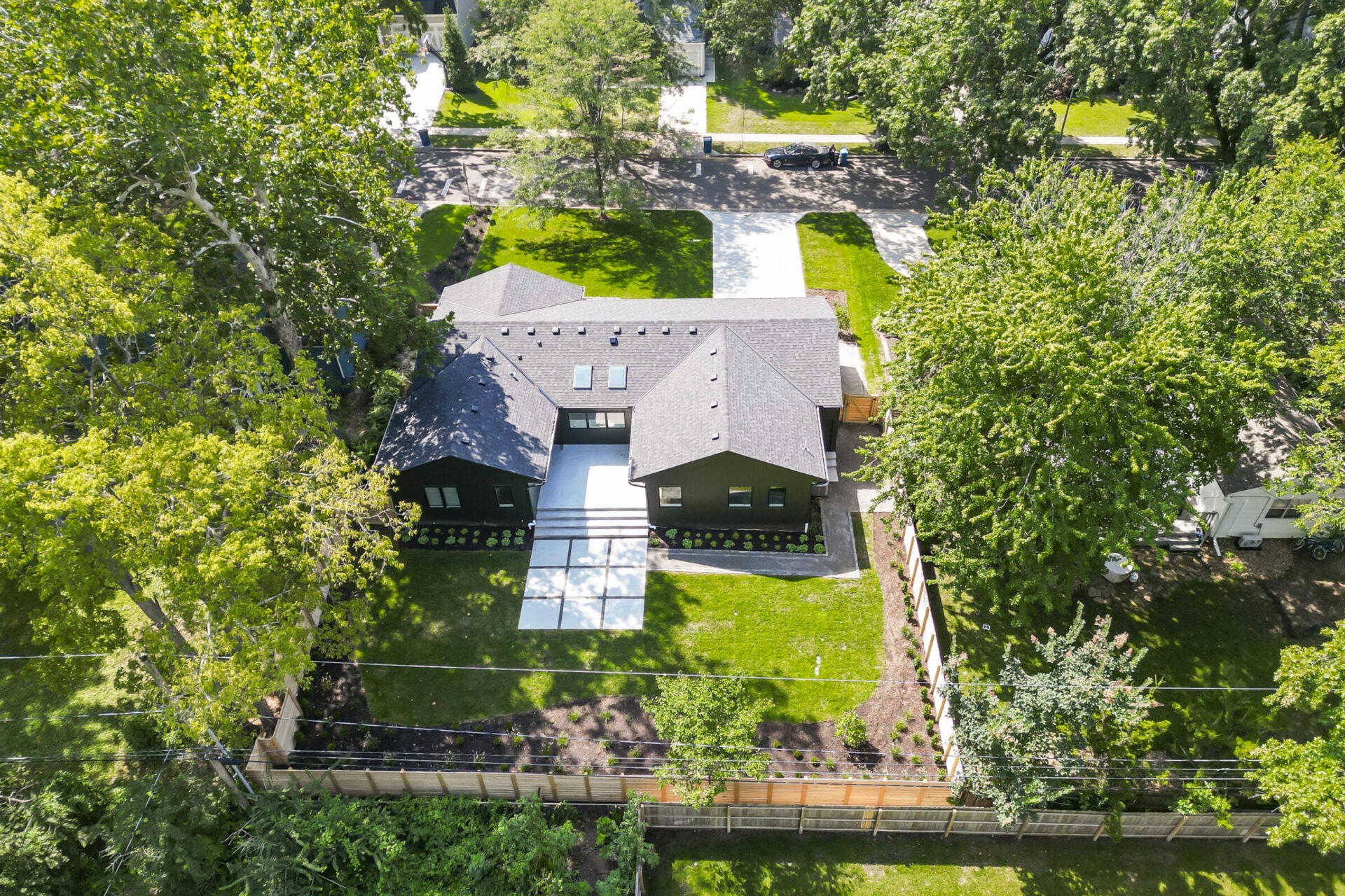 Aerial view of a modern house with a large yard, surrounded by trees and a wooden fence, with a driveway leading to the street.