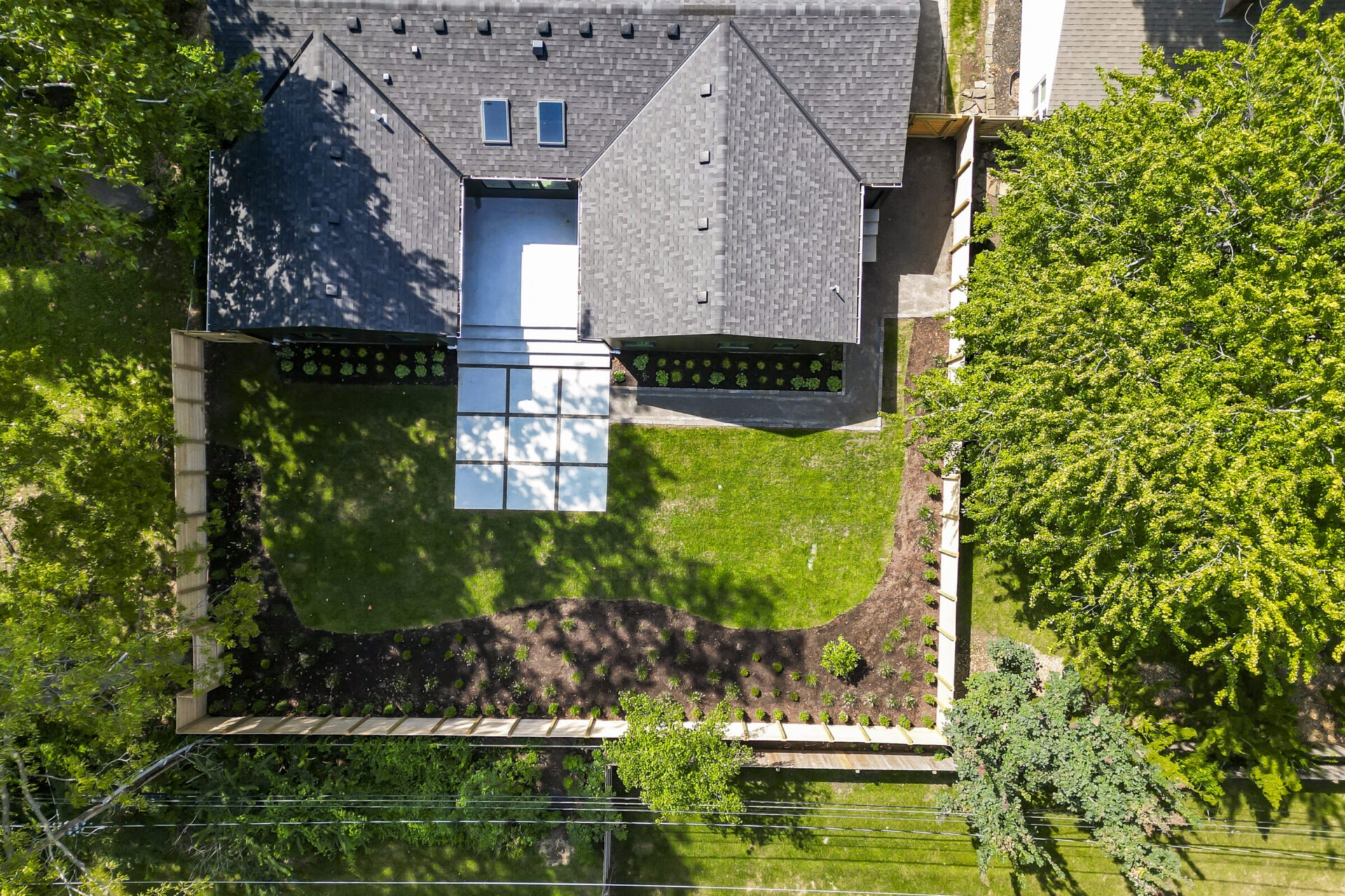 Aerial view of a modern house with a landscaped garden, fenced yard, and lush greenery surrounding the property, featuring a geometric patio design.