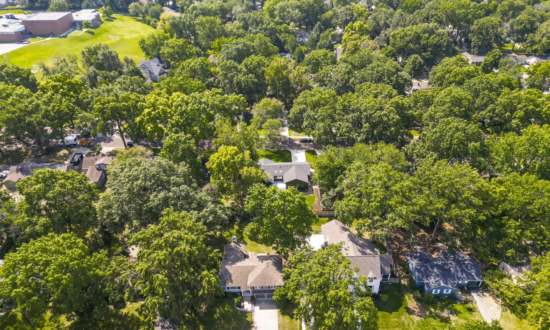 Aerial view of a suburban neighborhood with houses surrounded by lush trees, open green spaces, and a large building in the background.