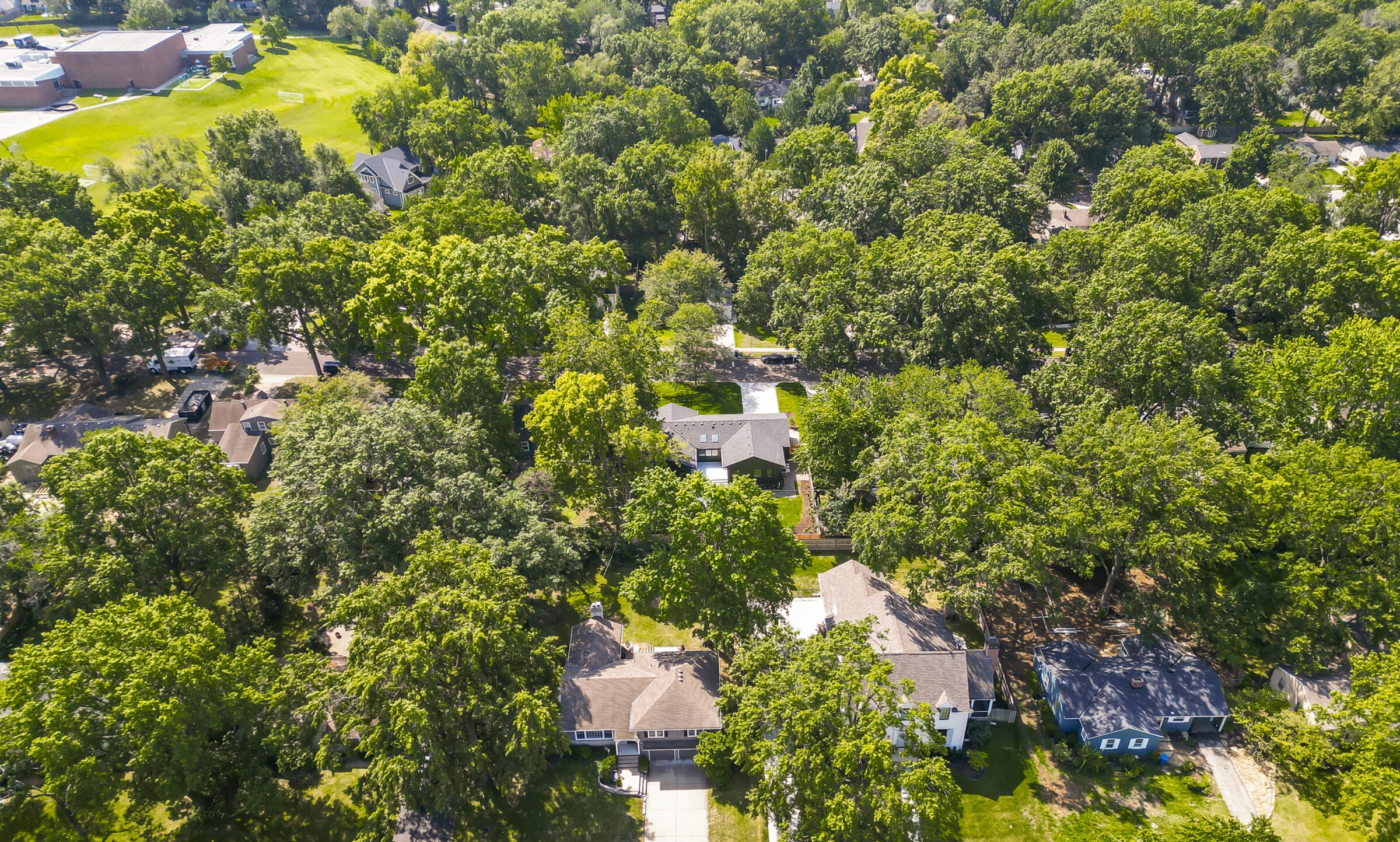 Aerial view of a suburban neighborhood with houses surrounded by lush trees, open green spaces, and a large building in the background.