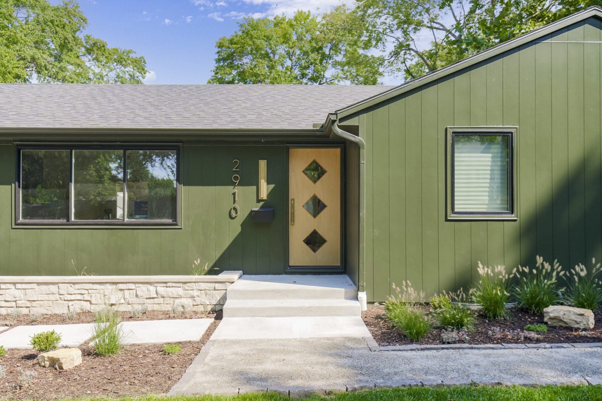 A green, single-story house with modern design, large windows, and a minimalist garden. The address number 2910 is by the door.