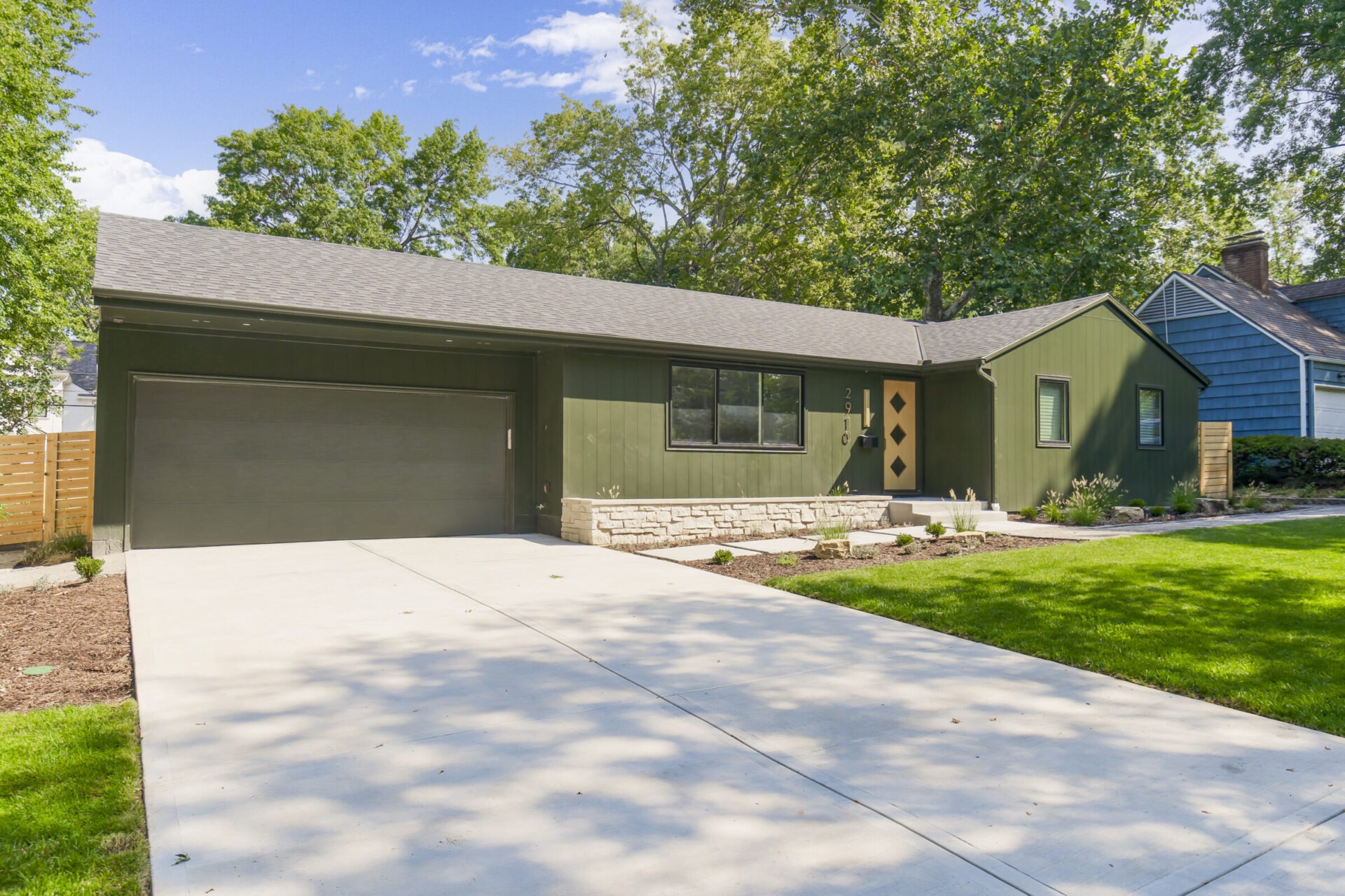 Single-story green house with attached garage, surrounded by trees and a well-maintained lawn. Clear driveway leads to main entrance. Sunny day.