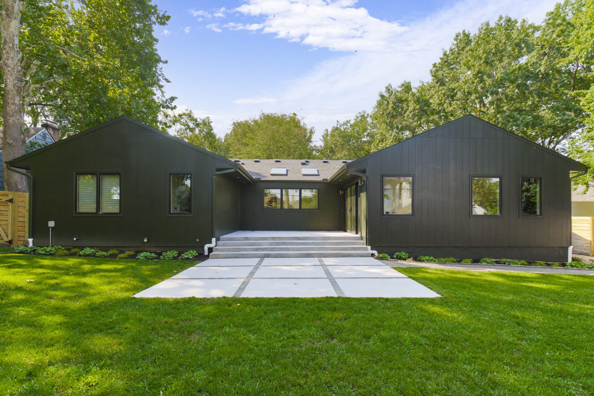 Modern black house with large windows, flat concrete walkway, and lush green lawn surrounded by tall trees under a partly cloudy blue sky.