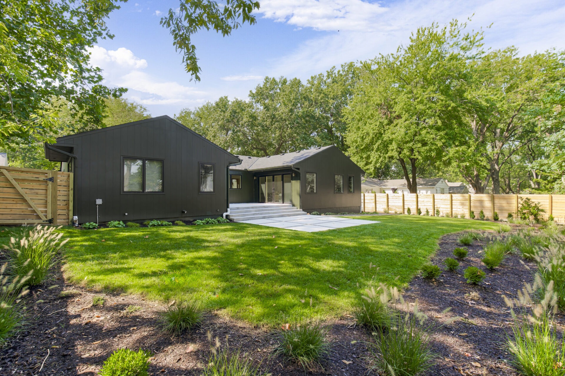 A modern black house with large windows, surrounded by a lush green lawn and wooden fence, set against a backdrop of trees.