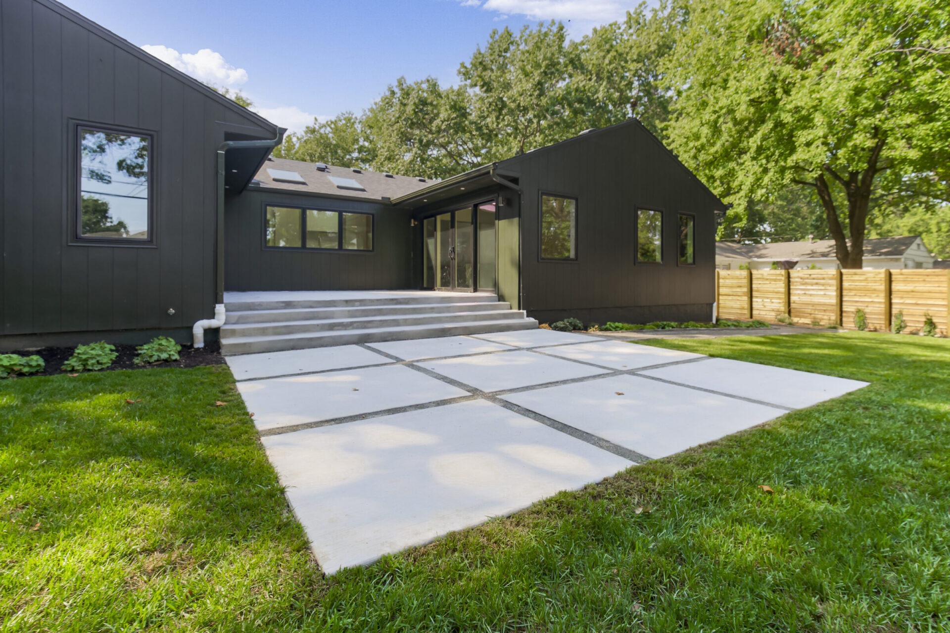 Contemporary black house with large windows and concrete steps. Spacious yard with green grass, trees, and wooden fence in a suburban setting.