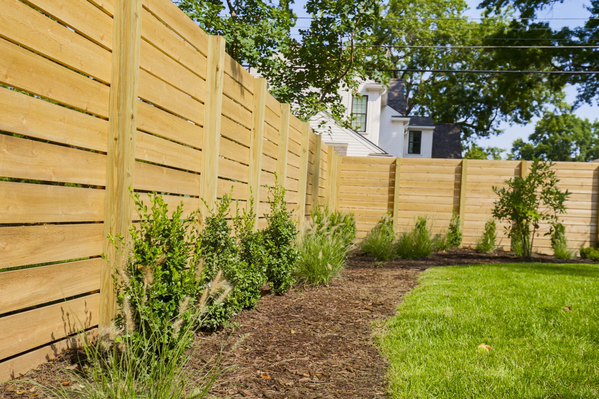 Wooden fence lines a garden with shrubs and grass, adjacent to a white house with a dark roof. Lush trees in background.