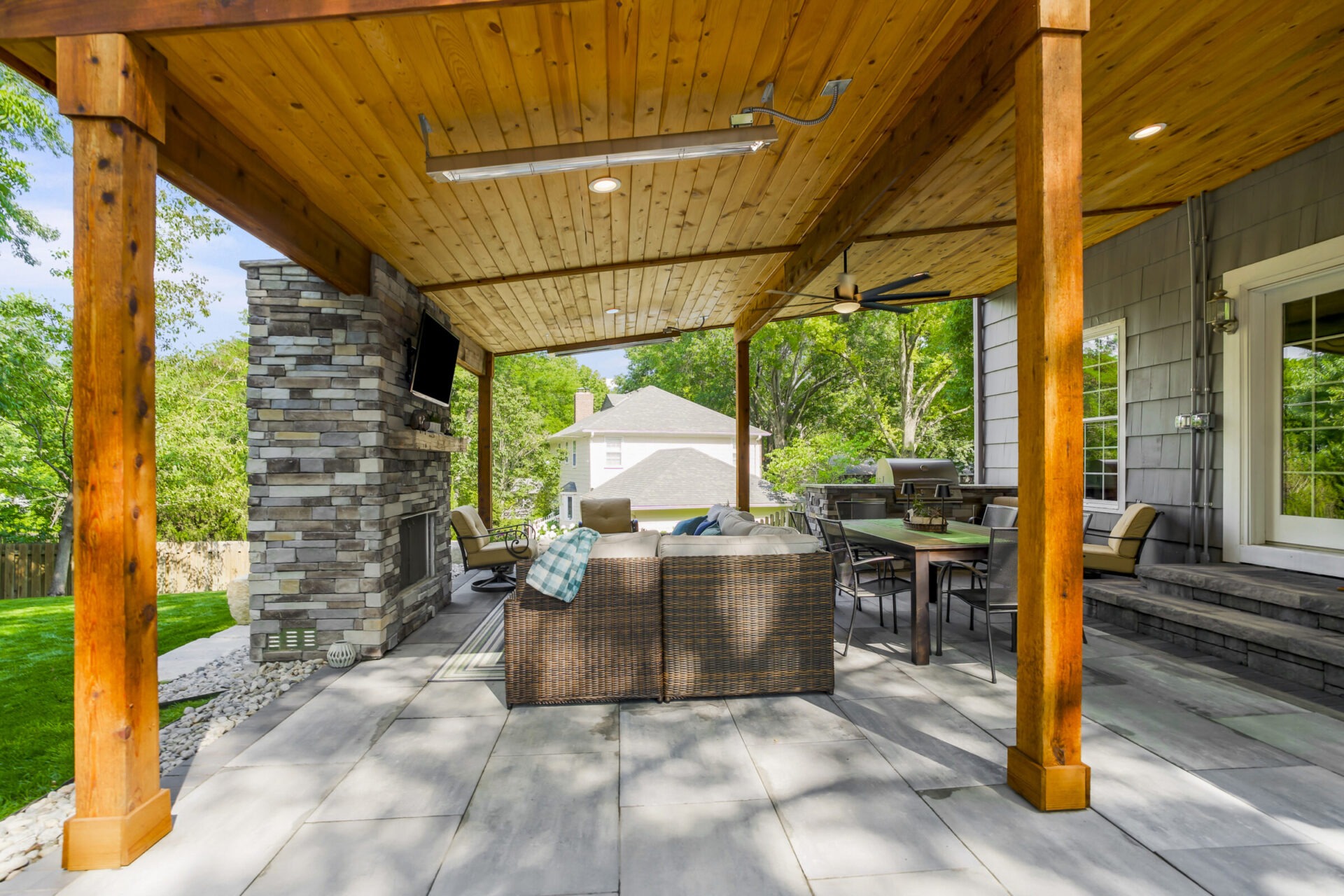 Covered patio with wooden ceiling, stone fireplace, and outdoor furniture. Overlooks a lush backyard and neighboring house. Peaceful and inviting outdoor space.