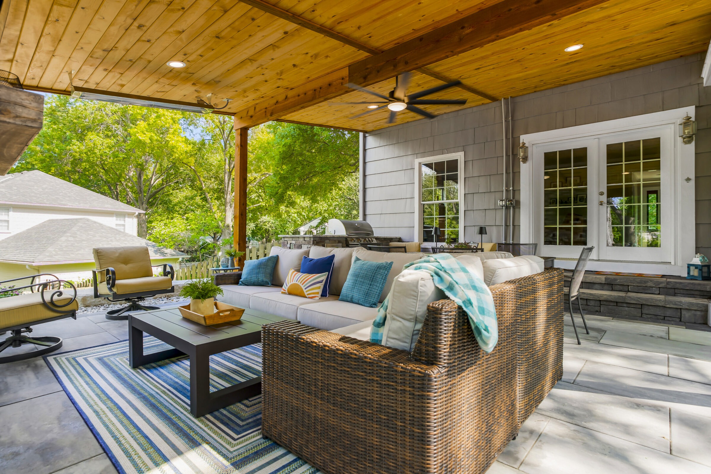 A cozy outdoor patio with wicker furniture, colorful cushions, and a wooden ceiling, surrounded by lush greenery on a sunny day.