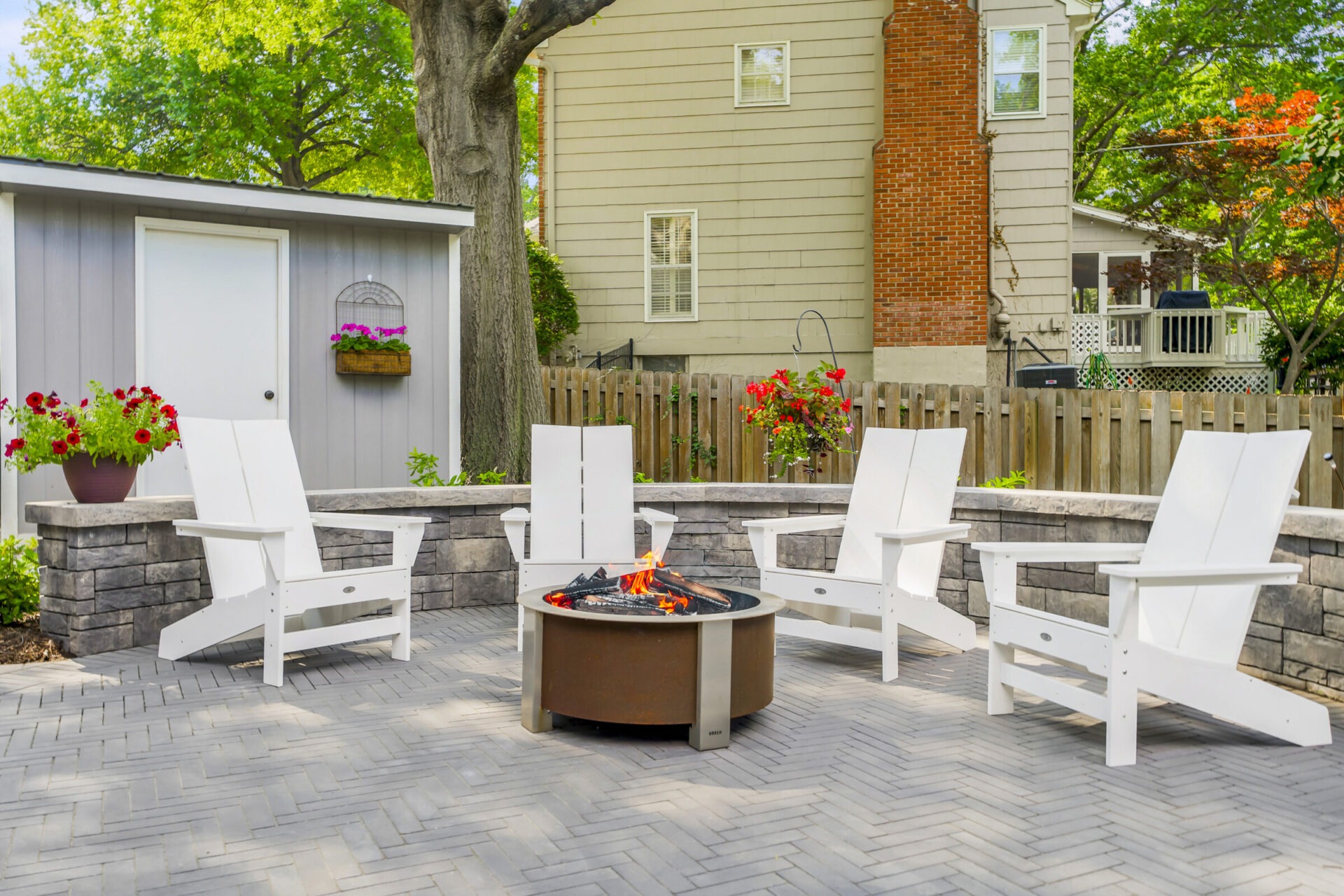 Four white chairs encircle a fire pit on a paved patio, surrounded by flowers, a tree, and a wooden fence.