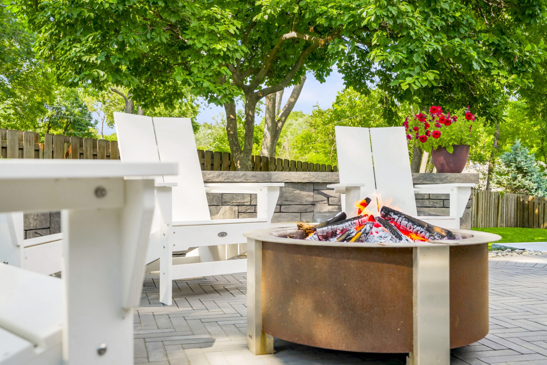 A cozy backyard scene with a fire pit, white chairs, vibrant flowers, and lush green trees under a clear blue sky.