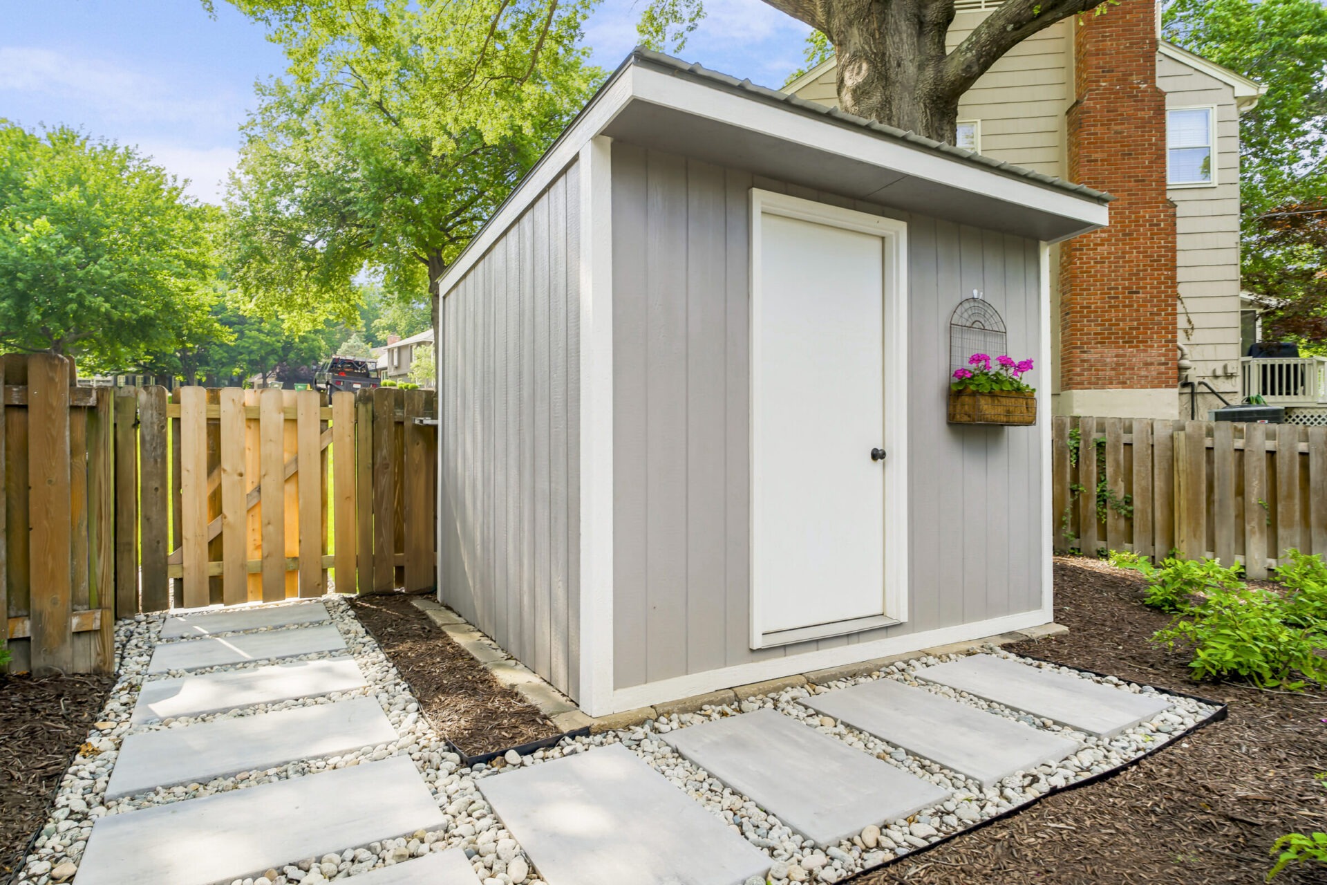 Small gray garden shed in a fenced yard, surrounded by trees and a brick house. Paved stone path and flower basket visible.