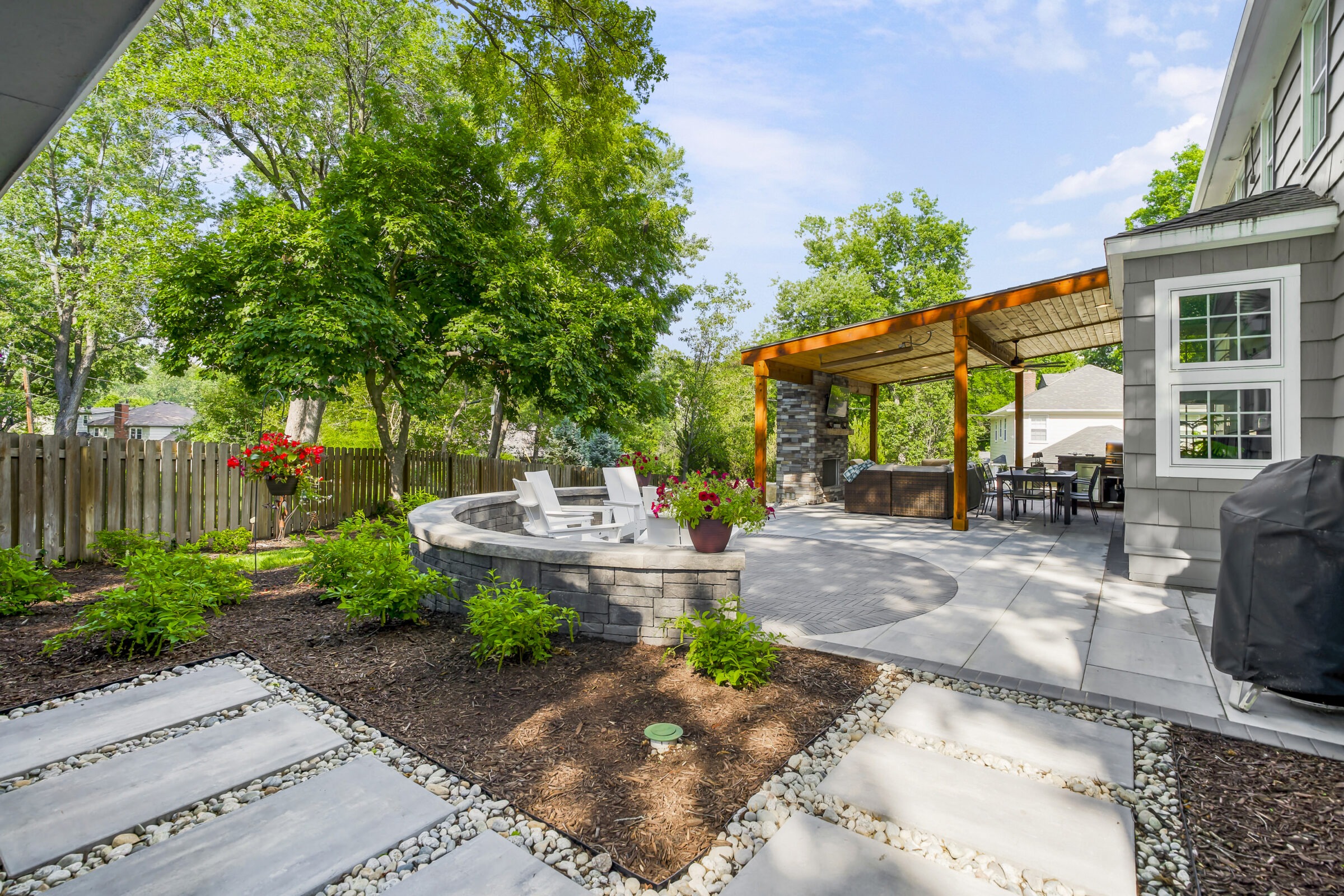 Charming backyard features a patio with white chairs, pergola, and greenery. Stone pathways lead through landscaped garden under a clear blue sky.