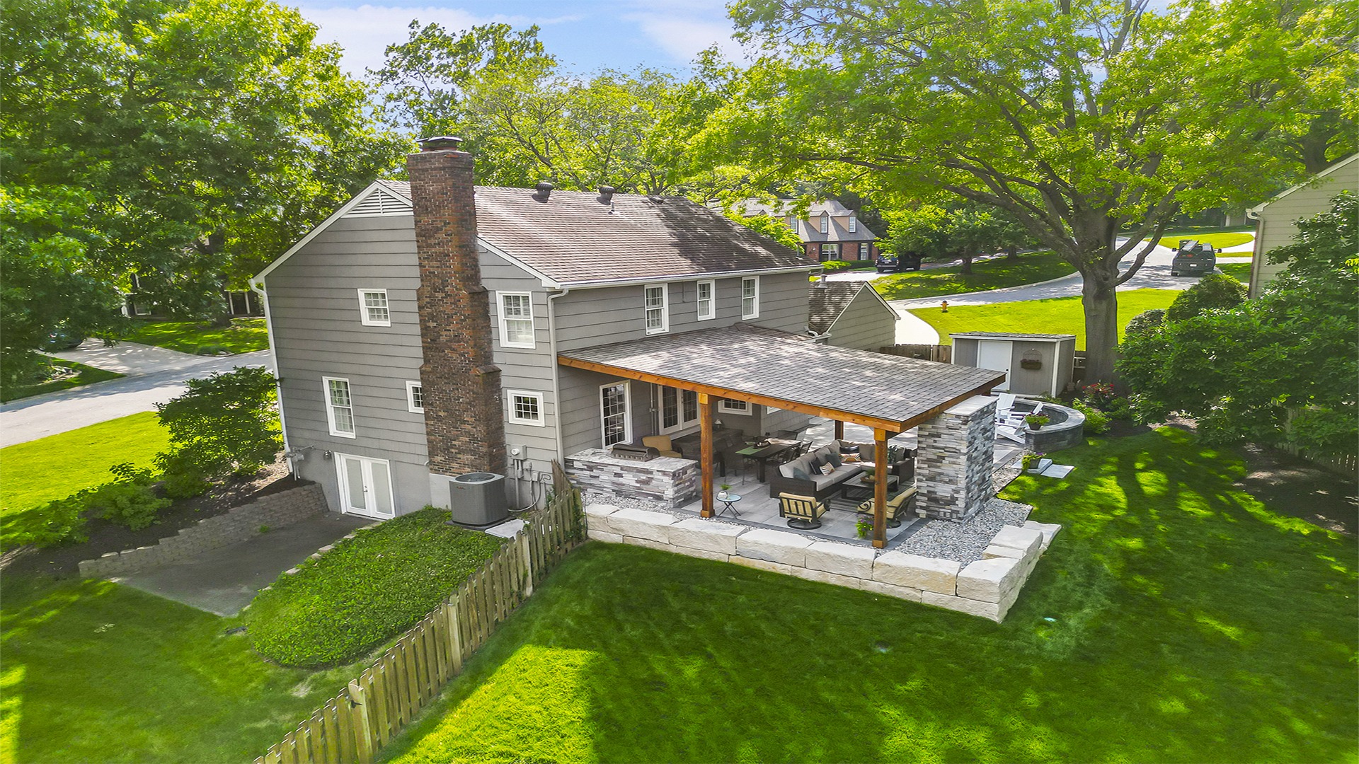 A gray house with a brick chimney features a covered patio and lush green yard, surrounded by trees on a sunny day.