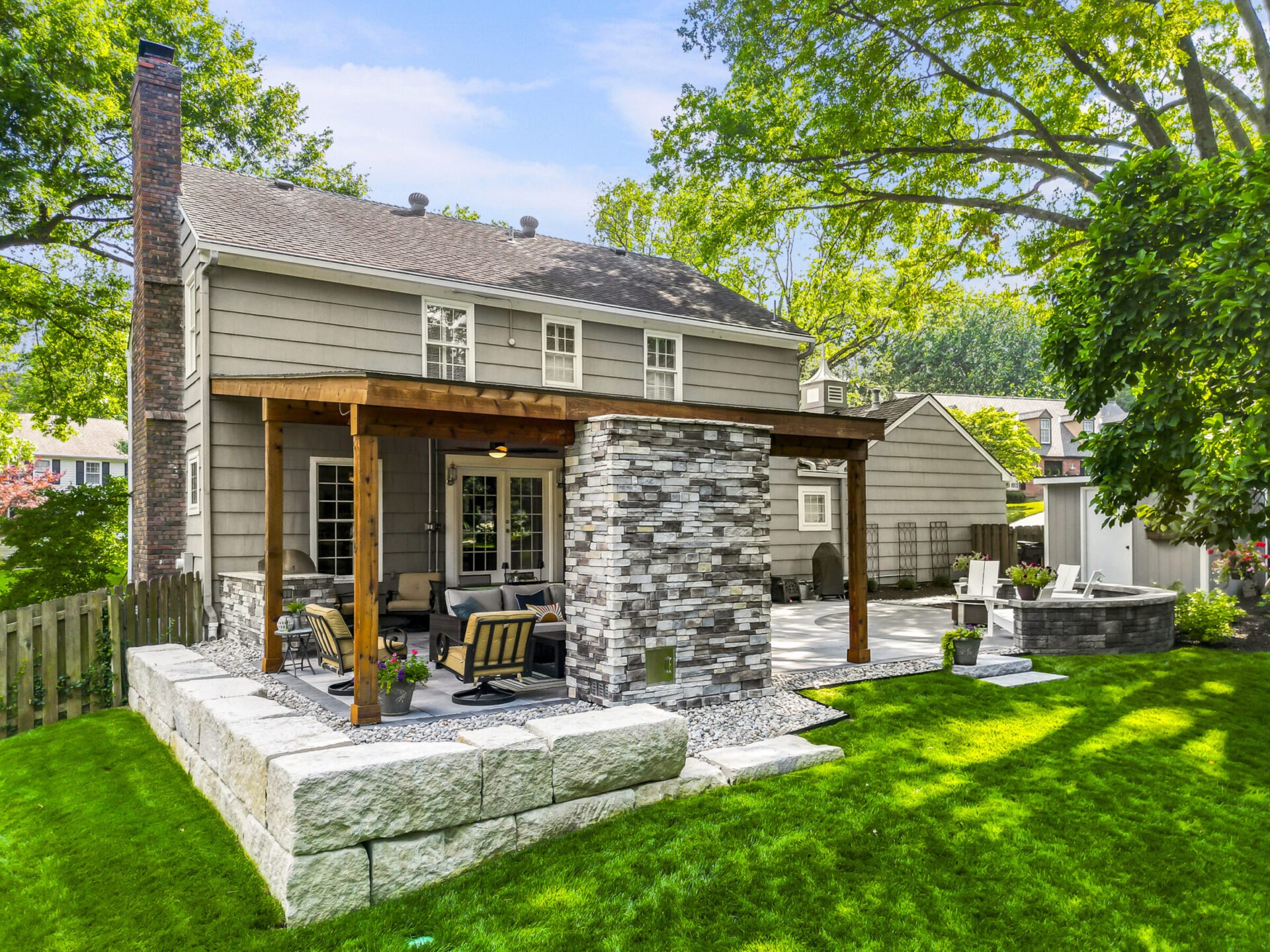 Suburban backyard with a stone patio, wooden pergola, and seating area. Lush green lawn and trees surround the gray house.