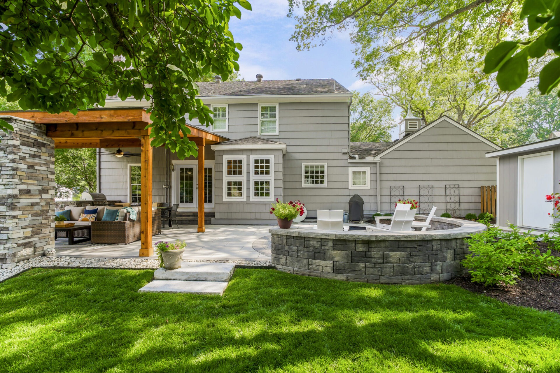 A charming backyard with a gray house, pergola, outdoor seating, lush grass, and a circular stone fire pit, surrounded by green trees.