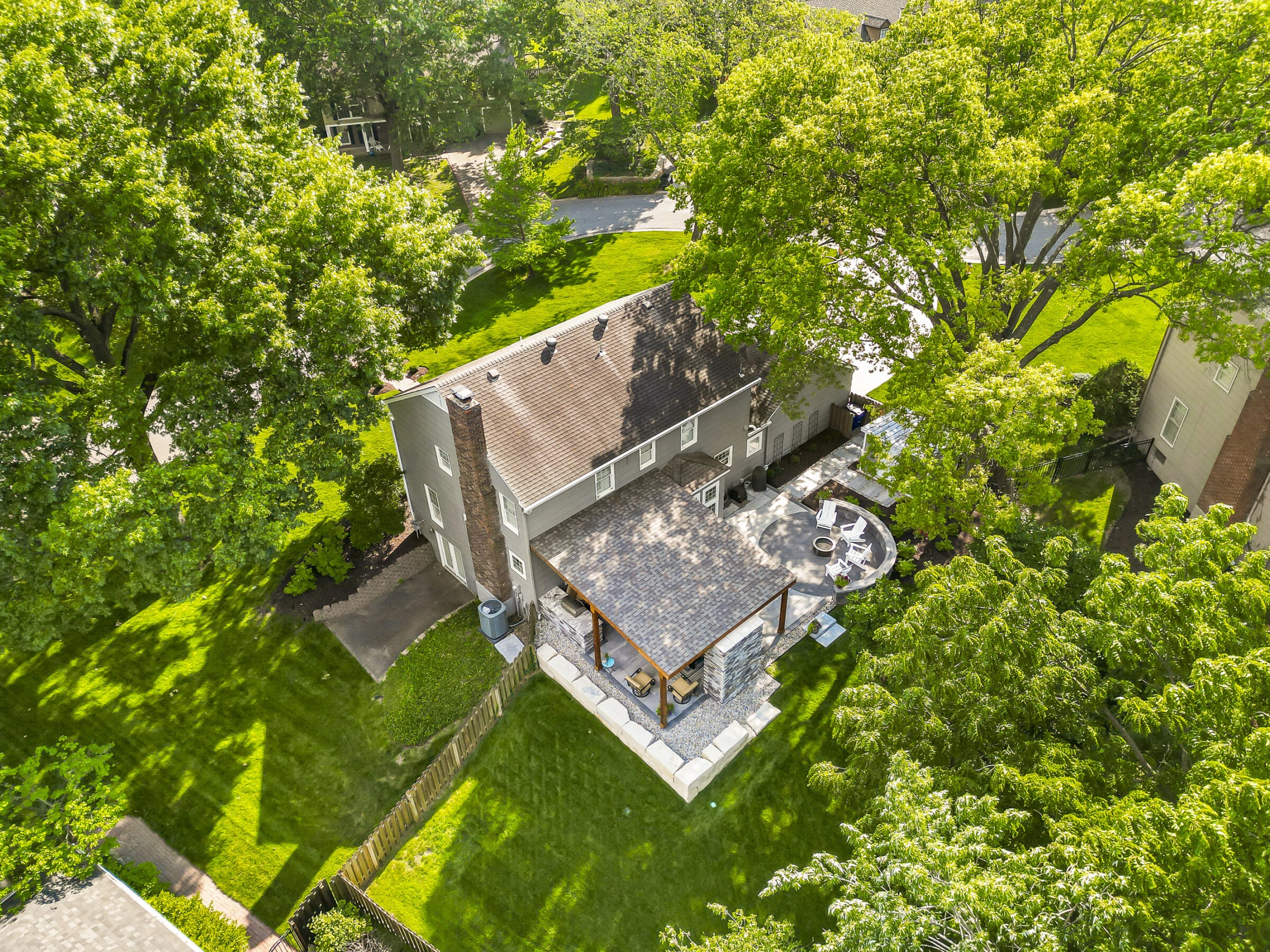 Aerial view of a suburban house with a spacious backyard patio, surrounded by lush green trees and well-maintained lawn in a residential area.
