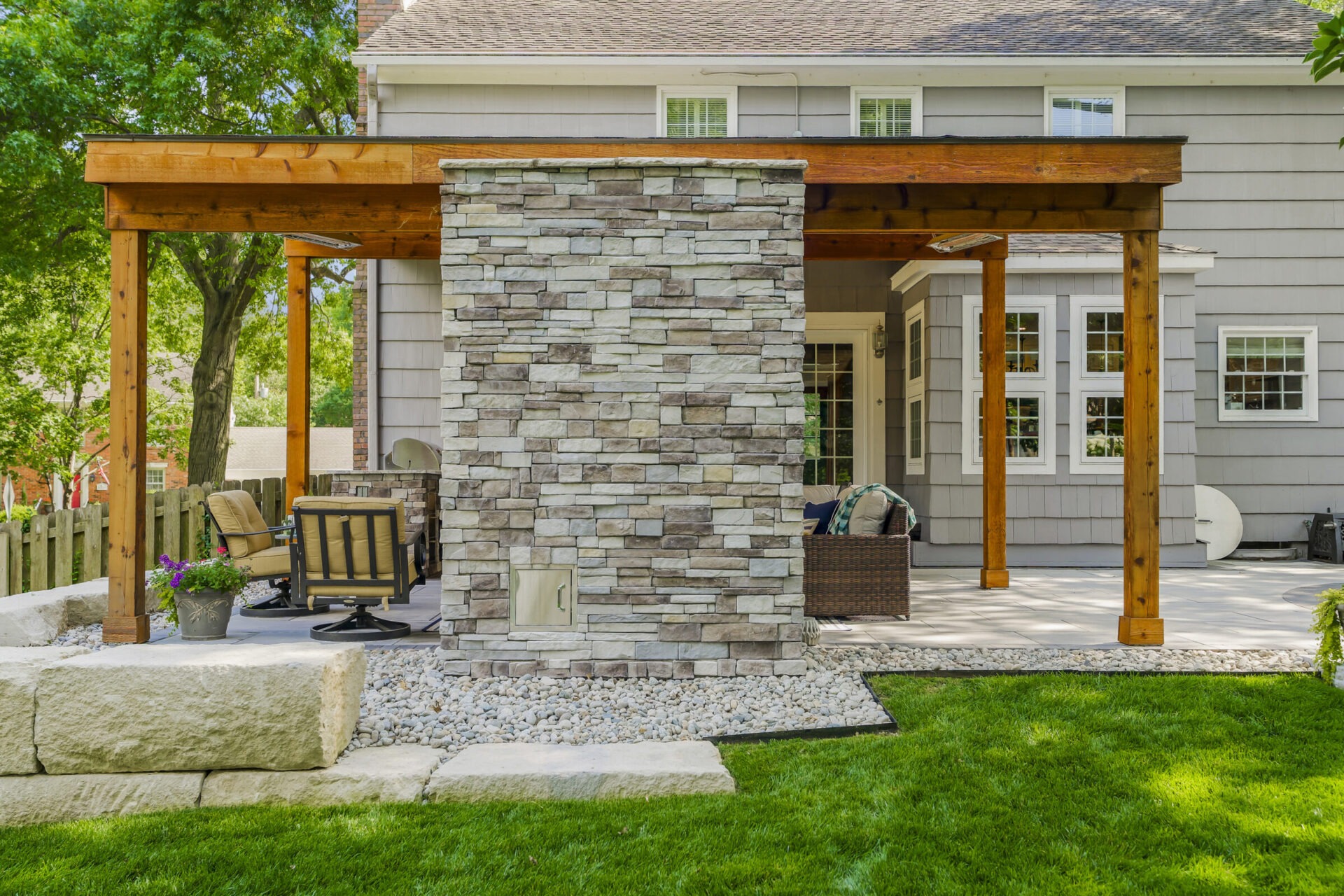 A backyard patio with wooden pergola, stone fireplace, and comfortable chairs. Lush lawn and surrounding trees create a tranquil outdoor setting.