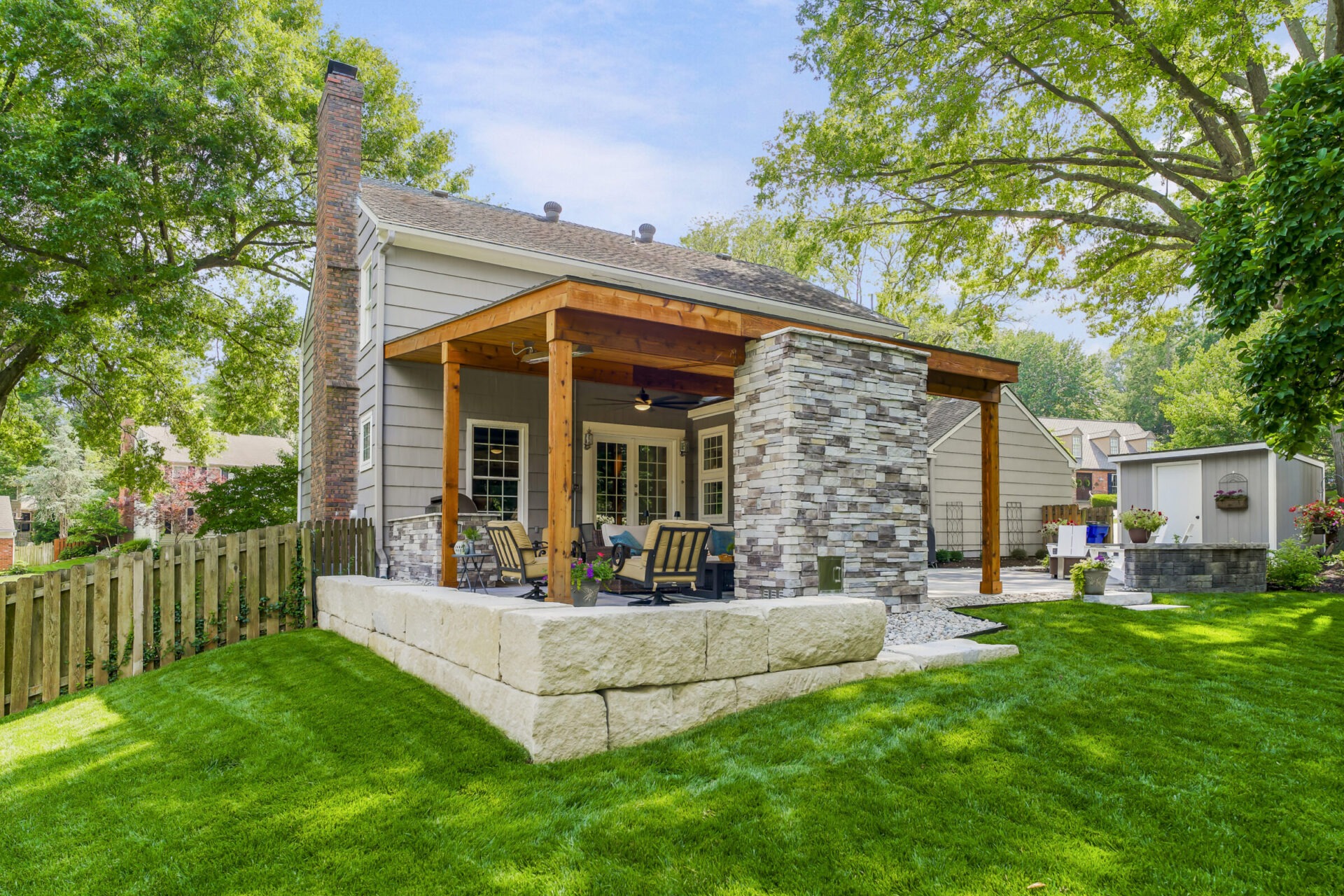 A house with a wooden pergola and stone patio, surrounded by lush green lawn and trees, features outdoor seating area.