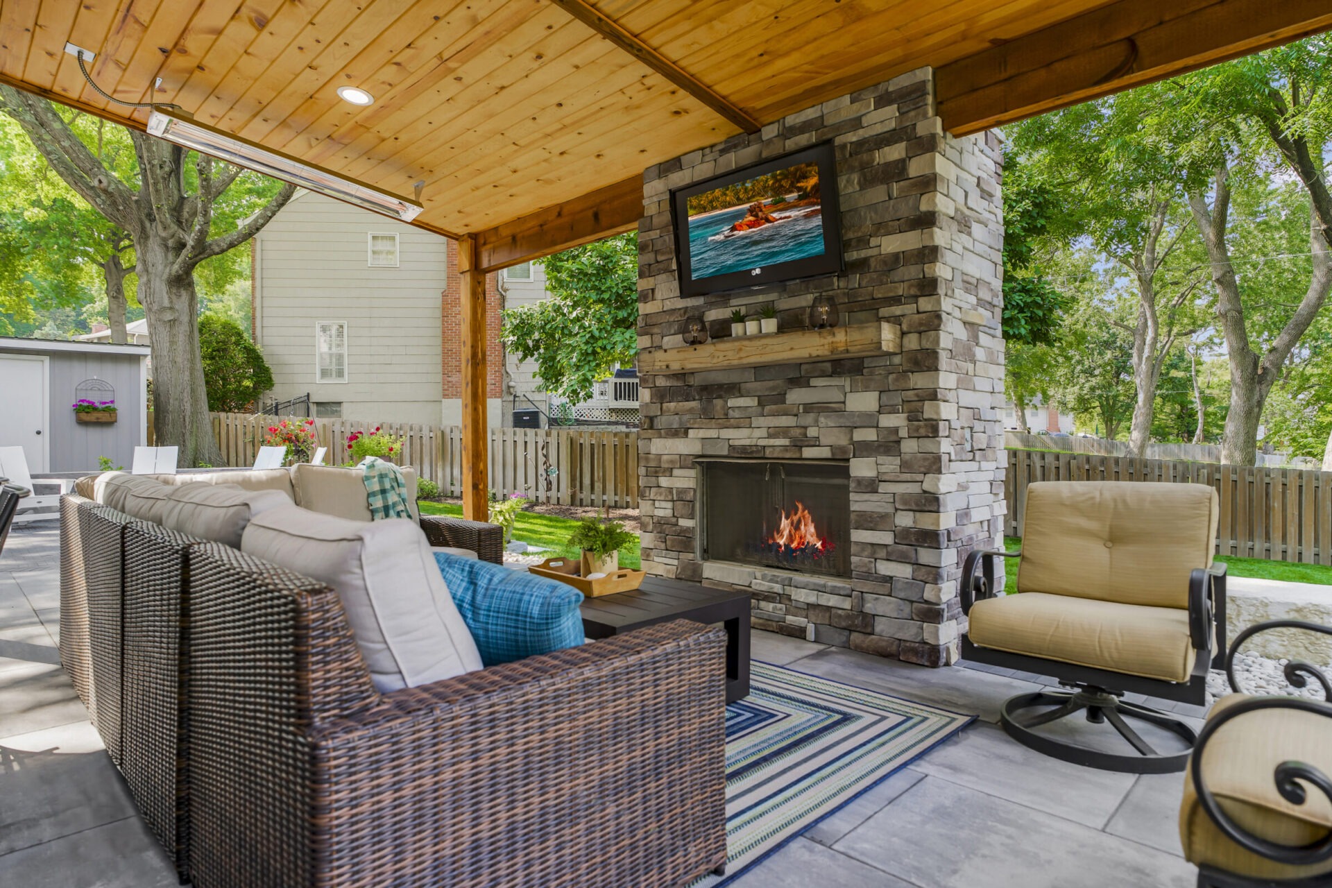 A cozy outdoor patio with wicker furniture, a stone fireplace, TV, and lush greenery. A house and trees are in the background.