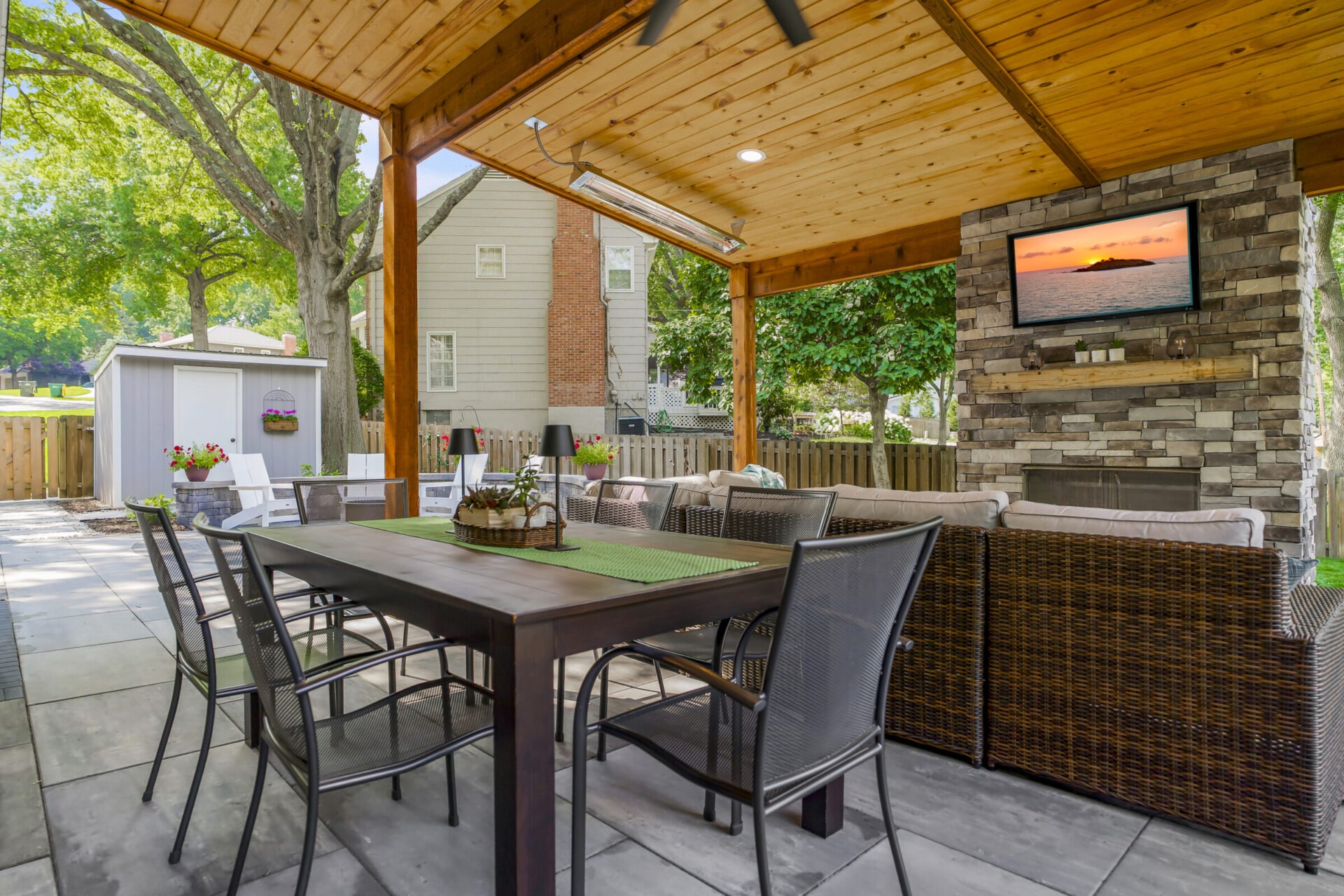 Covered patio featuring outdoor dining set, wicker furniture, and a television mounted on a stone fireplace. Surrounded by trees and residential fencing.