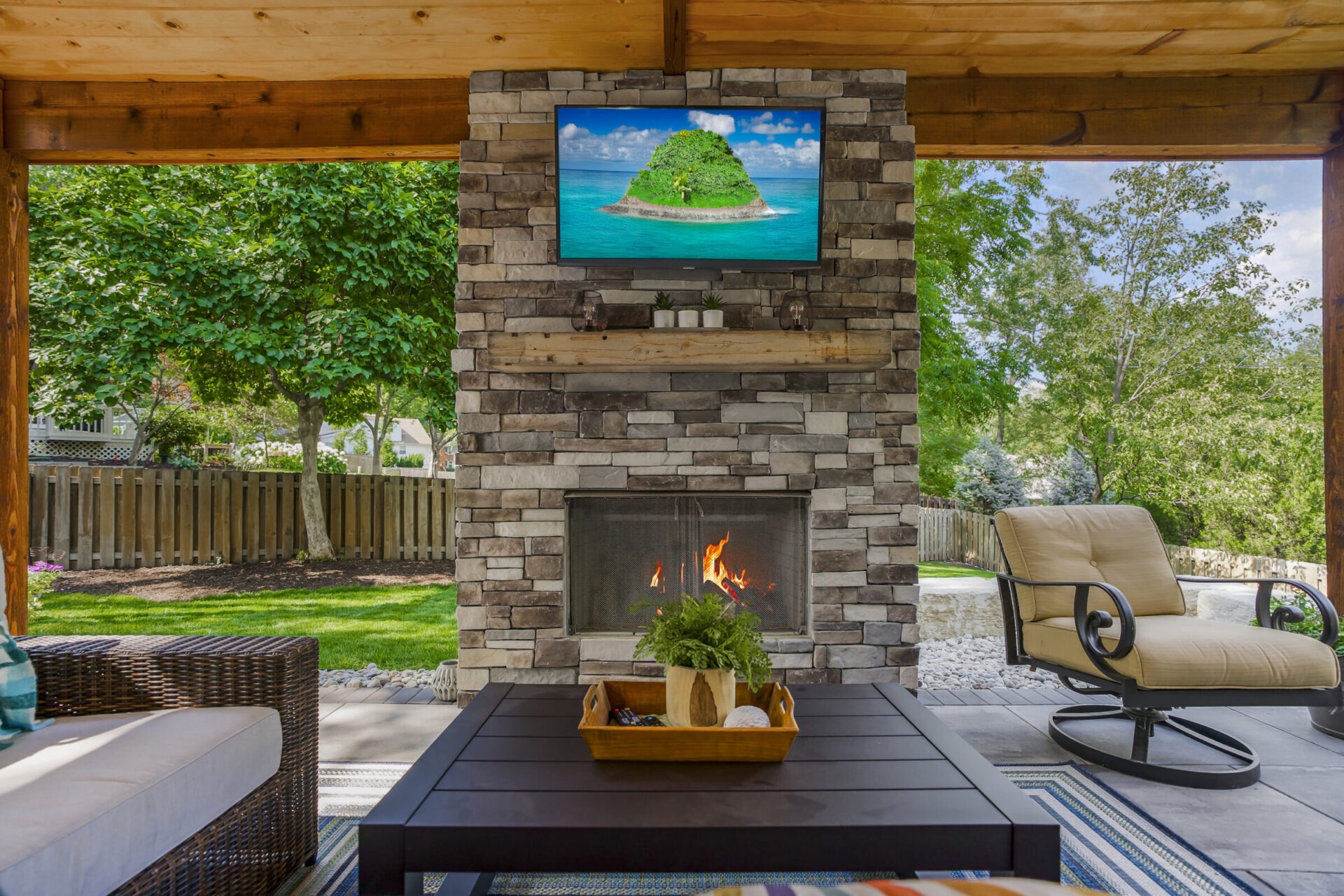 Outdoor patio with a stone fireplace, TV, and modern furniture. Green garden visible through wooden fence. Cozy and inviting atmosphere.