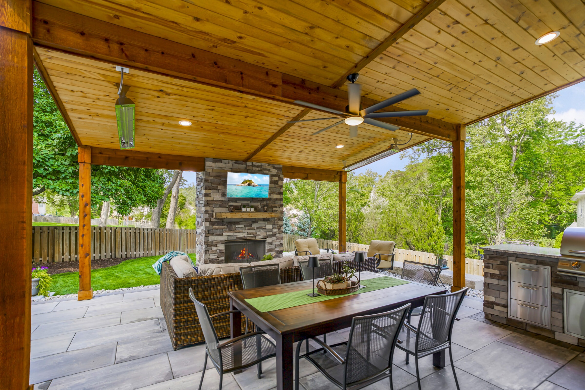 Covered patio with wooden ceiling, ceiling fan, outdoor fireplace, and dining table. Surrounded by trees and fenced backyard, comfortable seating arrangement.