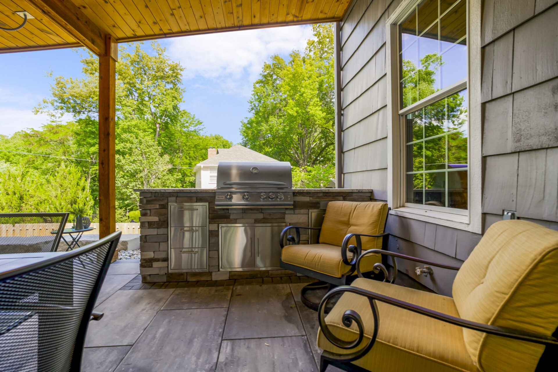 Covered patio with a built-in grill, two cushioned chairs, and outdoor table. Surrounded by trees and a neighboring house.