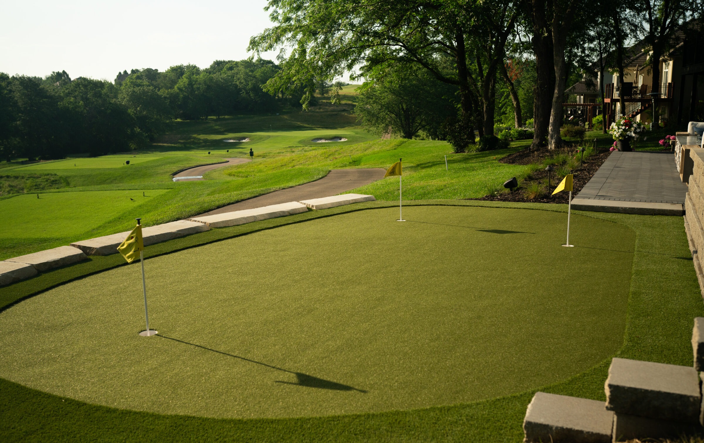 A scenic golf course with manicured greens and yellow flags, surrounded by lush trees and a paved path, under a clear sky.