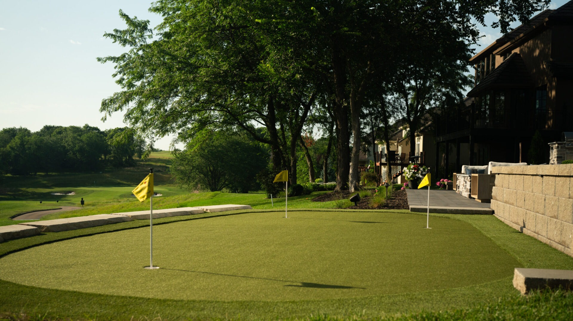 A tranquil golf course scene with putting green, surrounded by trees and houses. Yellow flags mark holes on the well-maintained grass.