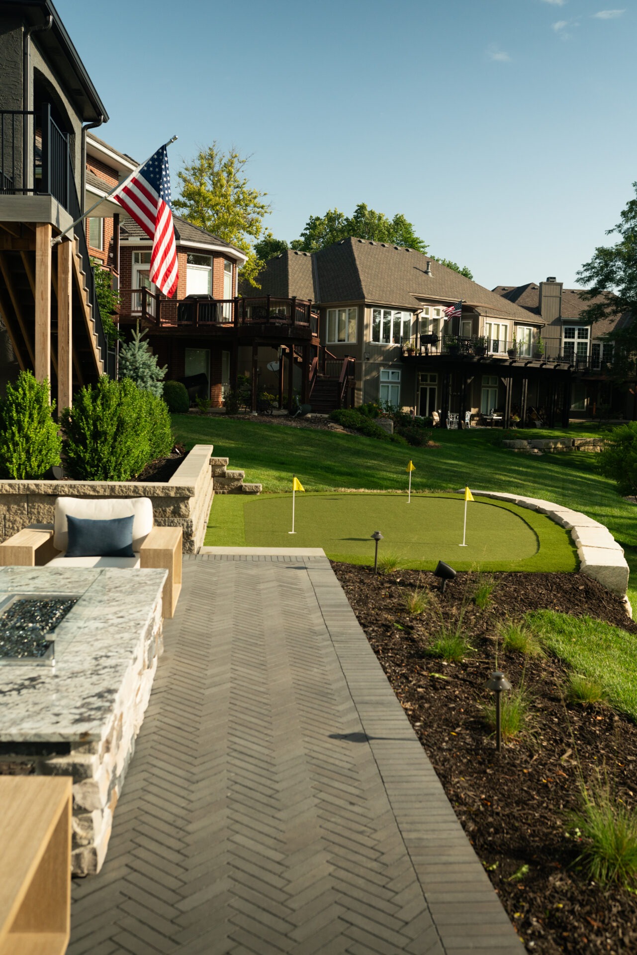 Suburban backyard with flag, cozy seating, and putting green surrounded by lush lawns, modern homes, and wooden decks under clear blue skies.