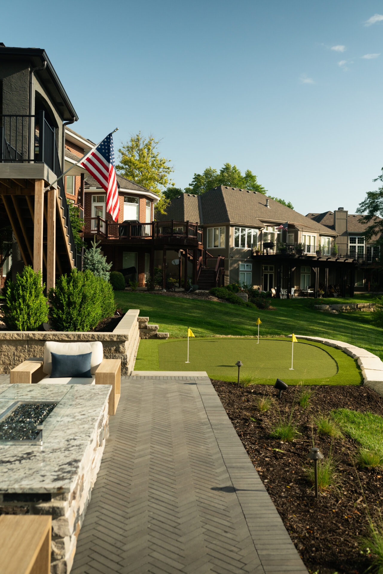 Modern backyard with putting green, patio, and American flag. Suburban houses with decks surround the landscaped area, under a clear blue sky.