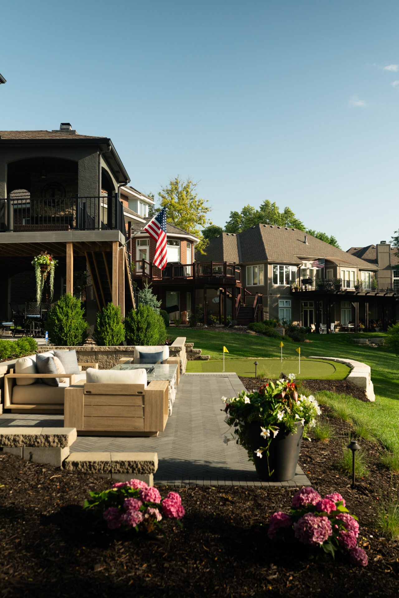 Spacious backyard with patio furniture, colorful flowers, and putting green. American flag displayed. Modern houses surrounded by lush greenery under a clear sky.