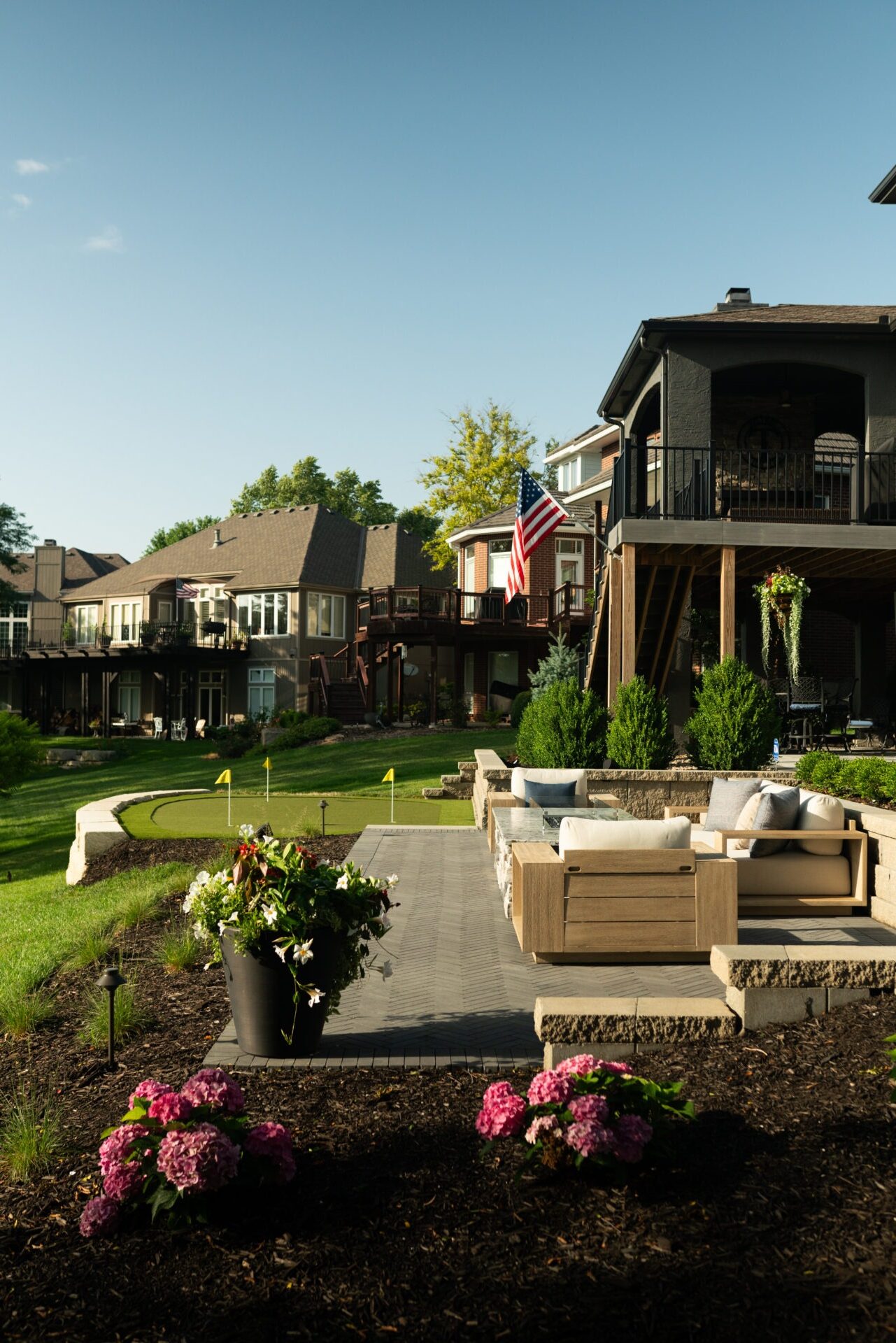 Spacious backyard with a cozy patio, lush garden, and small golf course. Houses feature an American flag, surrounded by greenery under clear skies.