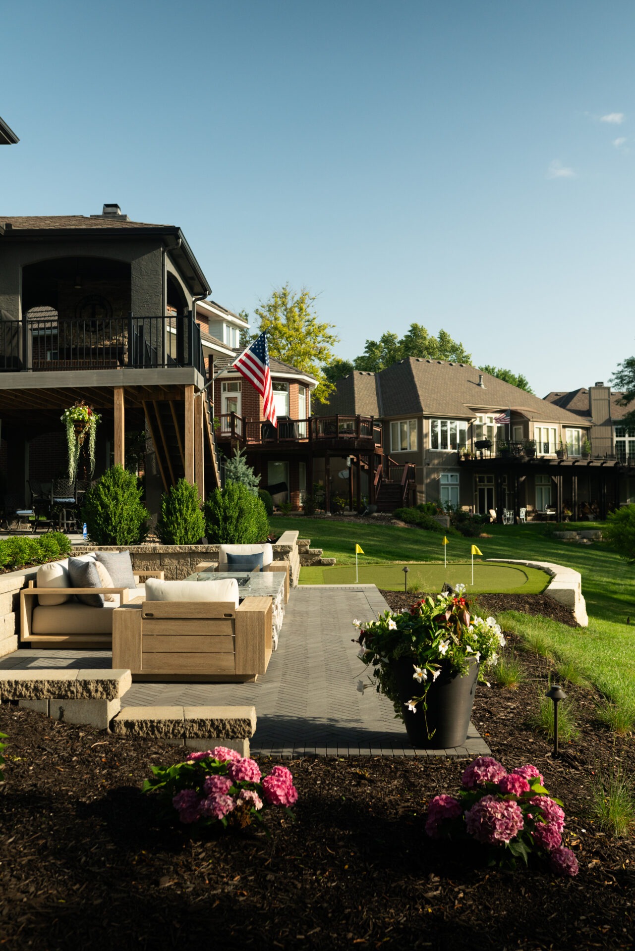 Beautiful backyard scene with patio seating, potted flowers, and an American flag. Residential homes surround a manicured lawn and putting green.