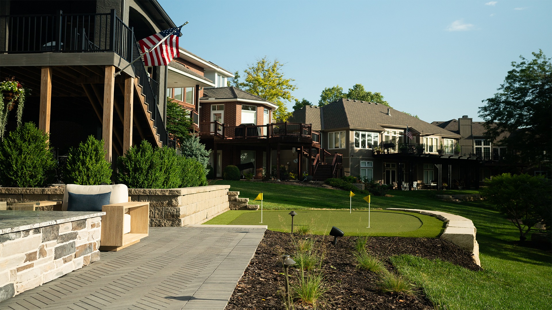 Suburban homes with spacious backyards, featuring a patio and a putting green. An American flag is displayed prominently. Lush greenery surrounds.