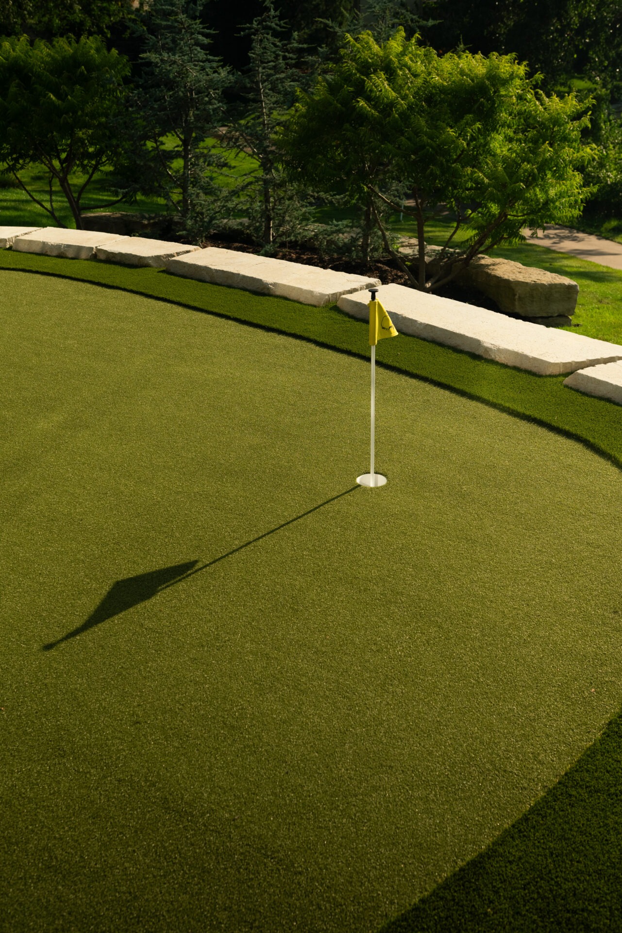 A lush, green putting green with a yellow flagstick, surrounded by trees and stone edging under clear, bright sunlight.