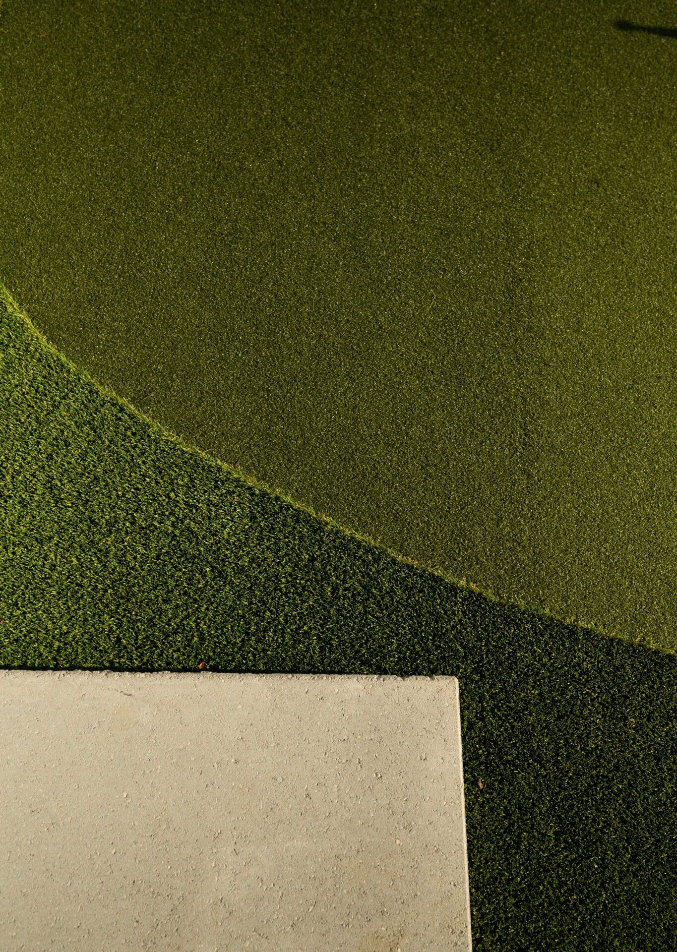 Artificial turf with varying shades of green forms abstract patterns, intersecting with a concrete path. Minimalist outdoor design, no landmarks present.