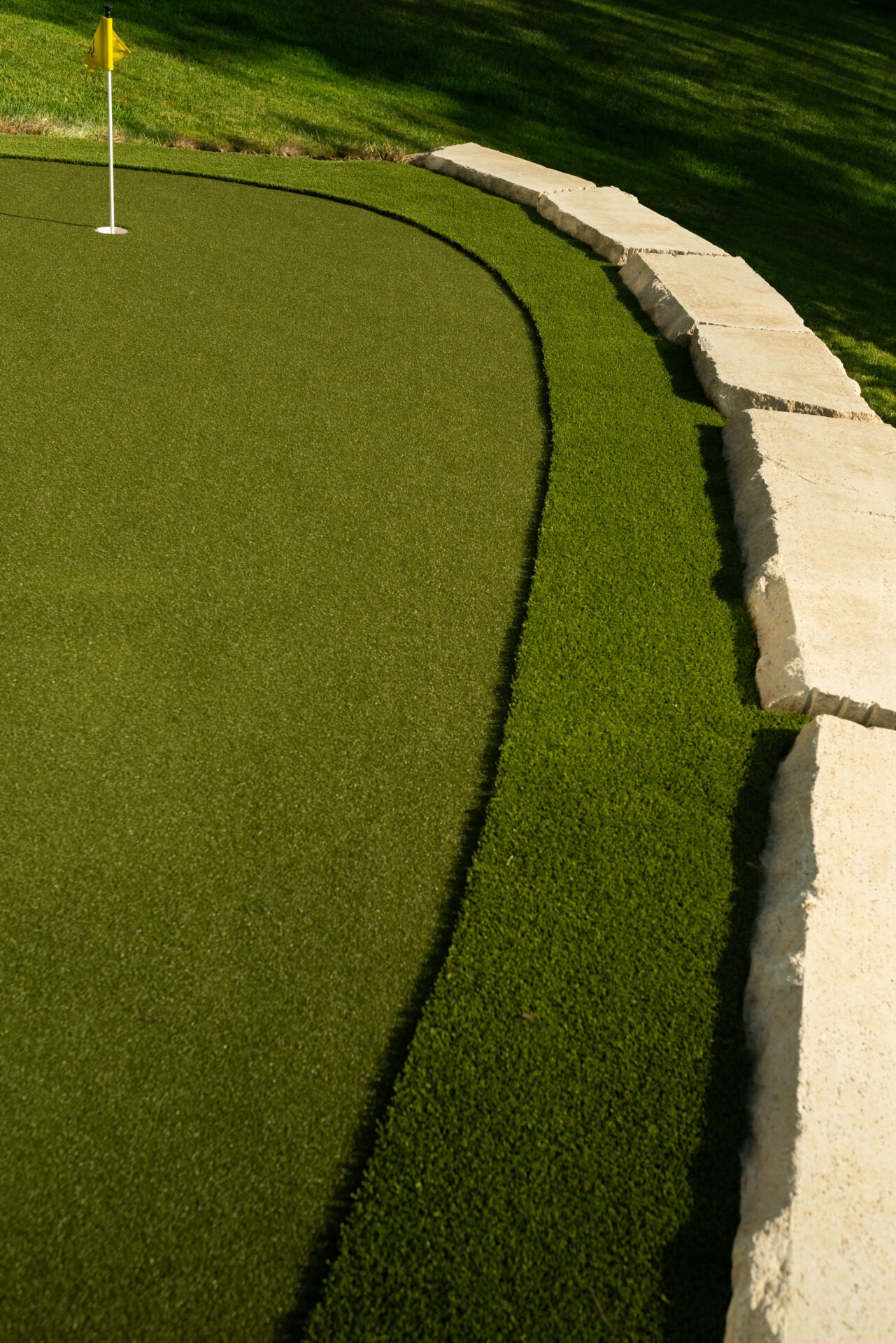 A putting green with a yellow flag, bordered by neatly aligned stone slabs under sunlight, showcasing lush artificial turf and grass.