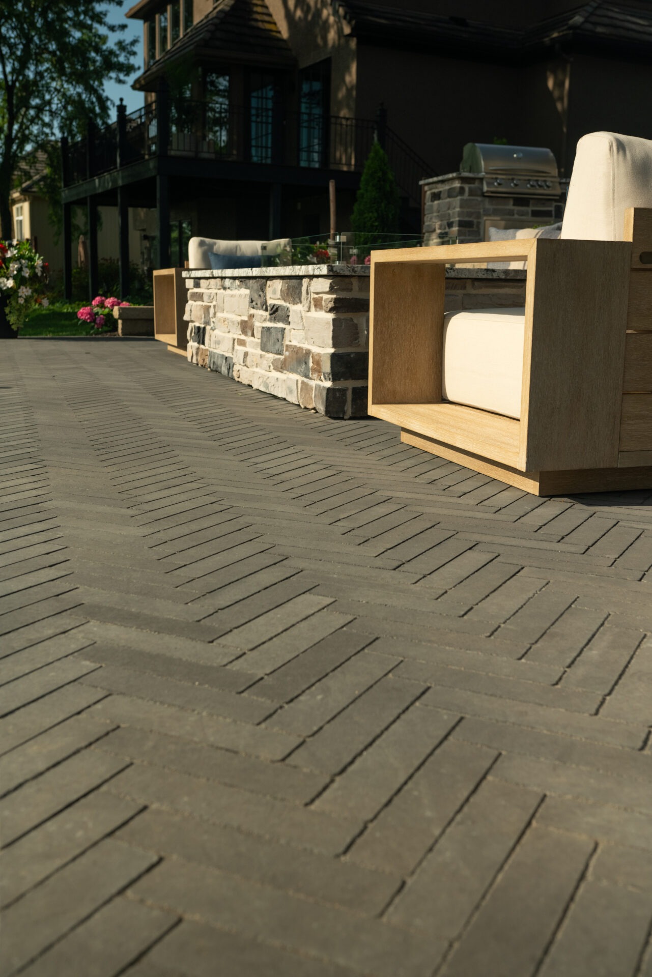 Stone patio with herringbone pattern leading to a modern house, featuring outdoor seating and a grill, surrounded by lush greenery and flowers.