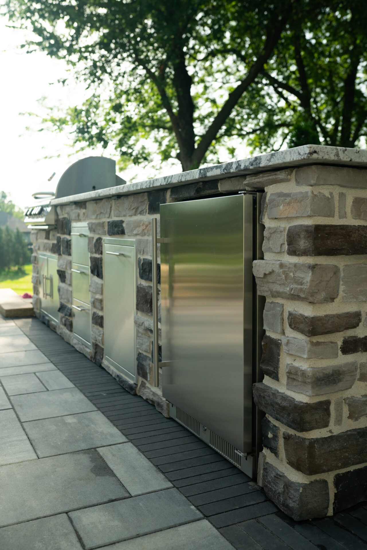 Stone outdoor kitchen with stainless steel appliances beneath shady trees on a sunny day.