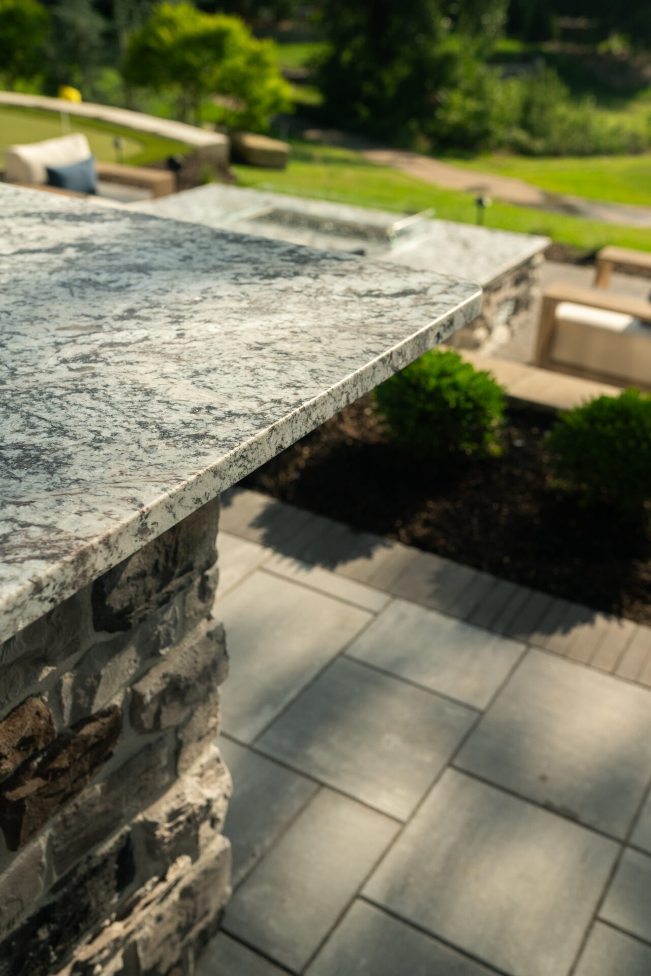 Stone patio with granite countertop, outdoor furniture, and neatly trimmed greenery. Sunlit garden setting with lush grass and shaded pathway in view.