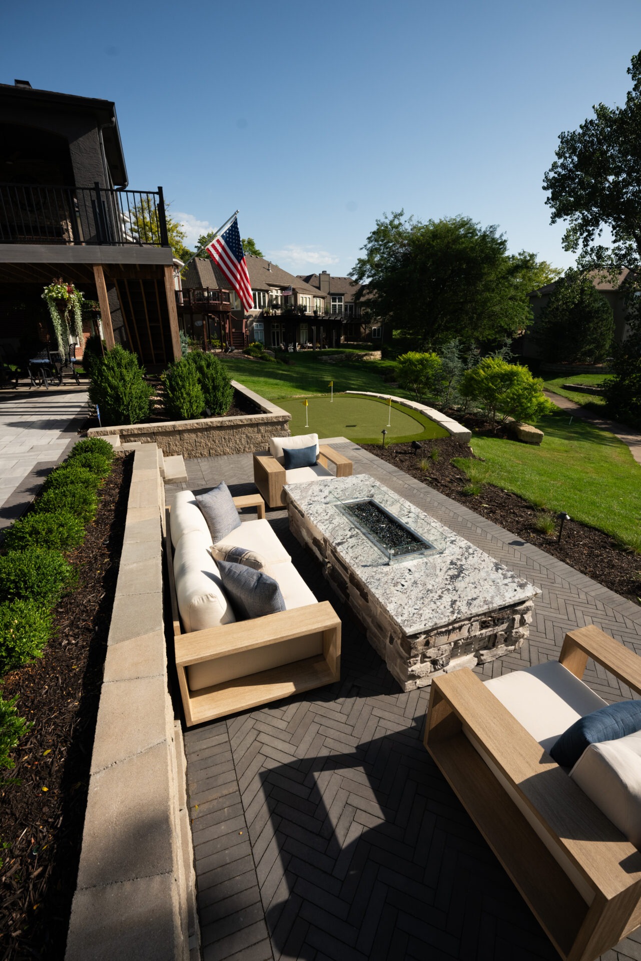 Patio with modern seating, fire pit, and a putting green. American flag visible, surrounded by lush greenery and neighboring houses under a clear blue sky.