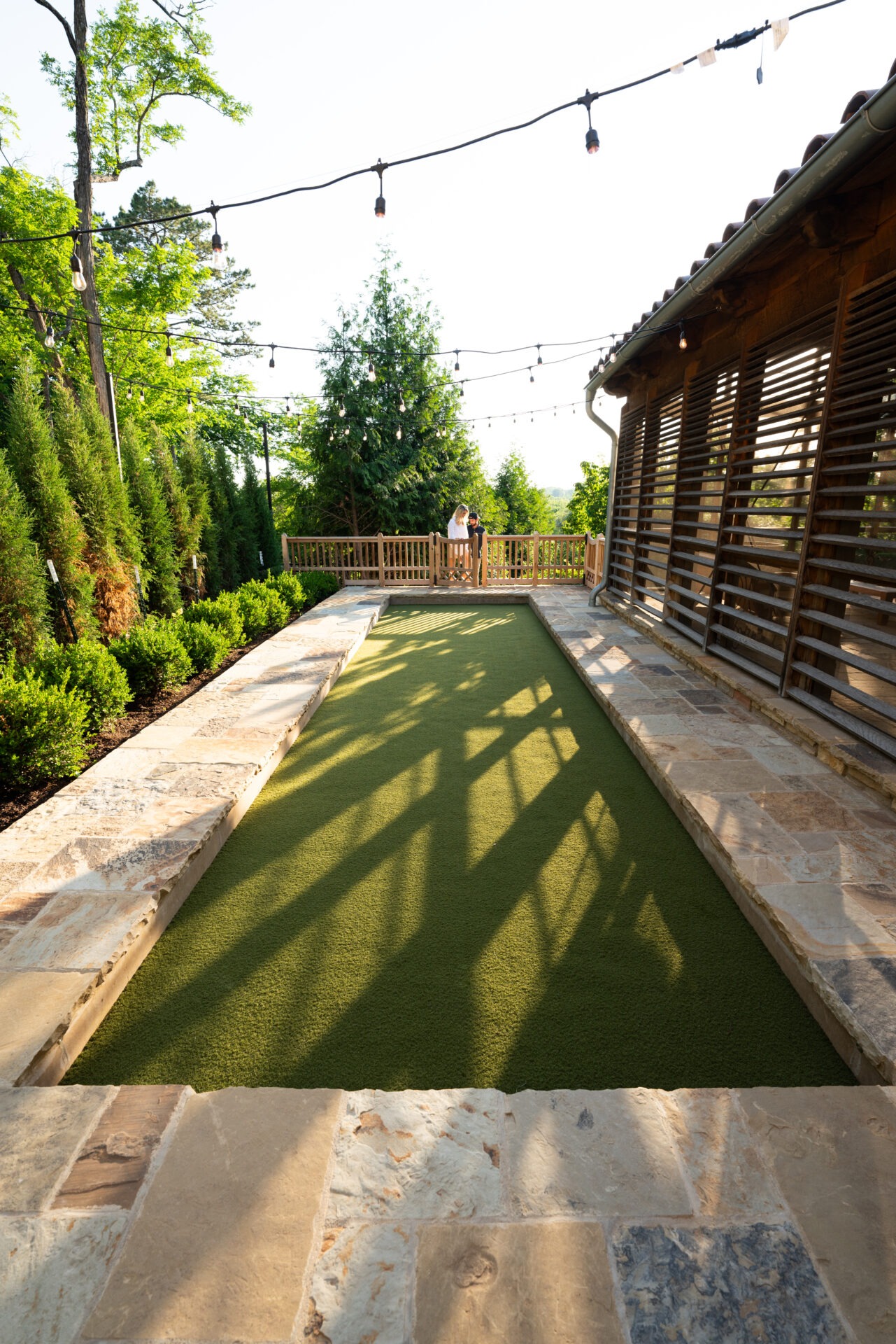 A person stands on a sunlit patio near trees, string lights, and wooden fencing, beside a bocce court surrounded by stone walls.