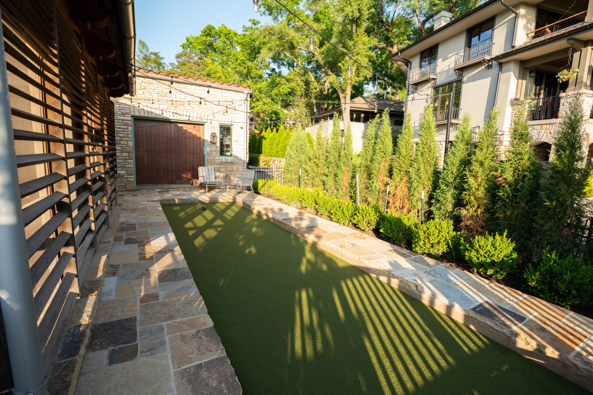 A stone patio with string lights and hedges surrounds a narrow green lawn, adjacent to modern houses and lush trees.