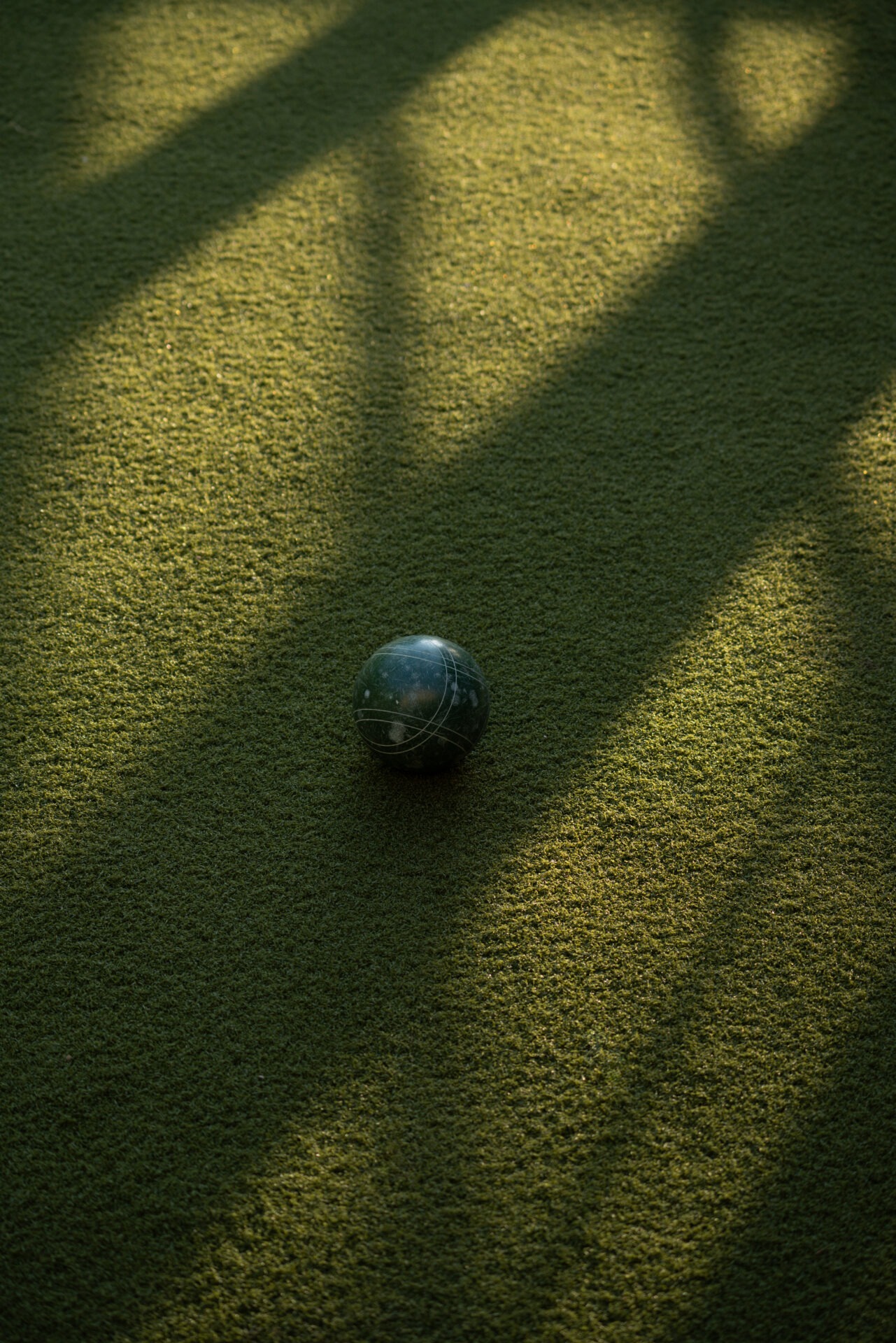 A single green ball rests on a textured surface with long shadows stretching diagonally in soft, warm lighting.